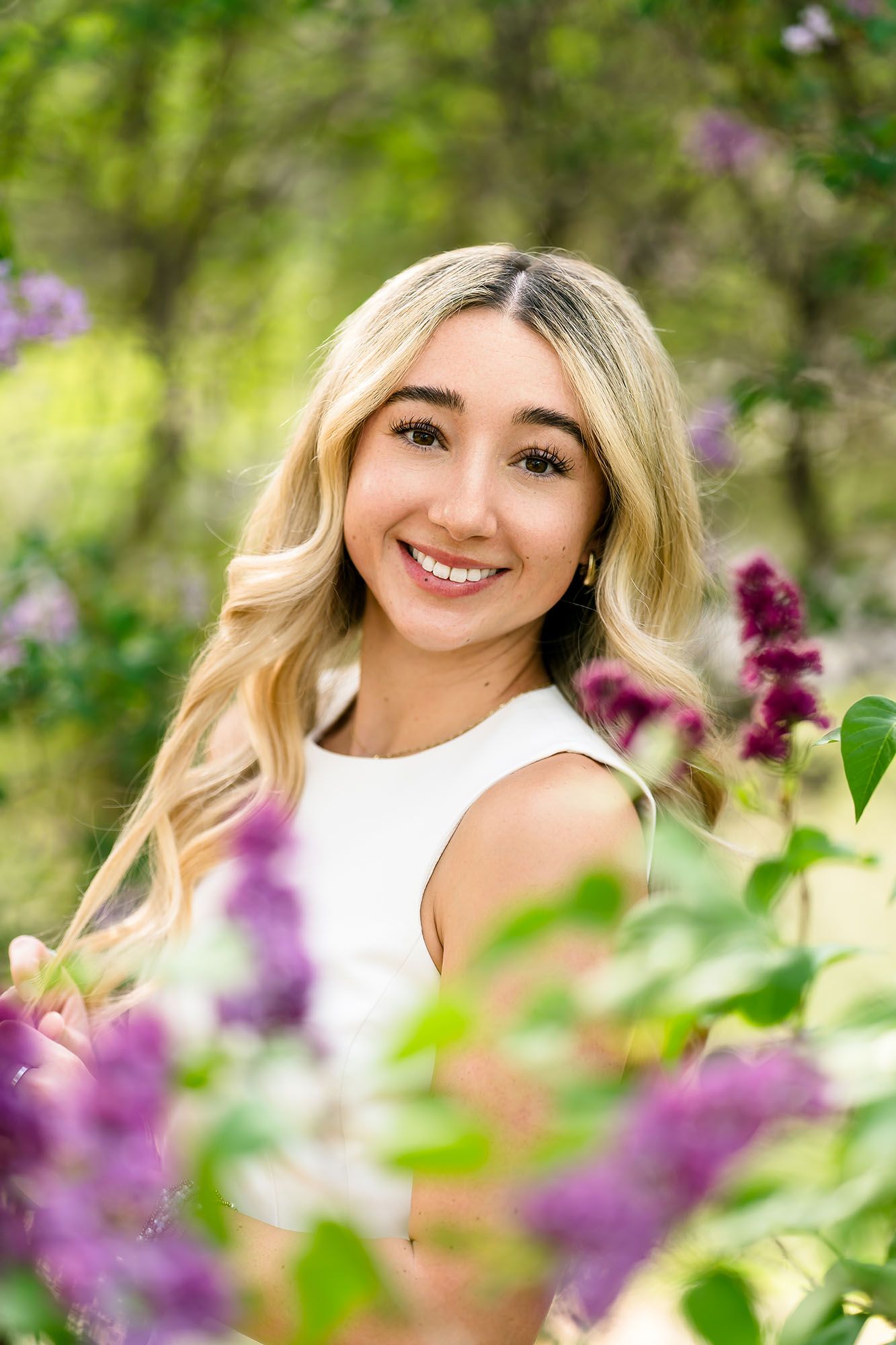 Smiling blonde teen girl surrounded by purple lilac blooms in lush green garden setting.