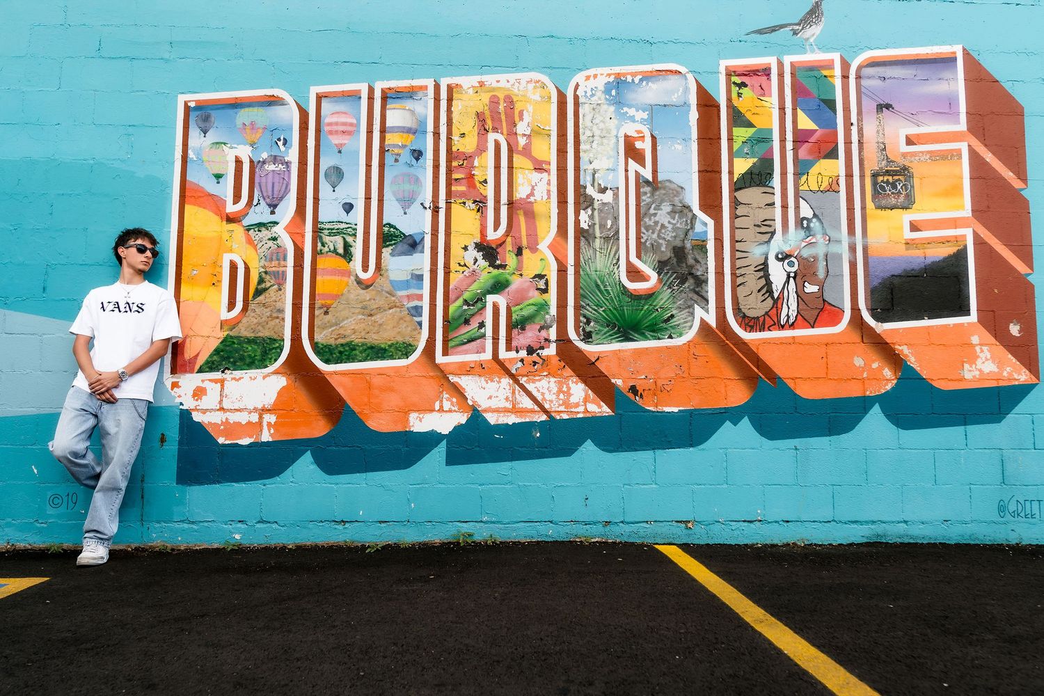 Young man posing in front of colorful Burque street mural on teal wall in urban setting.