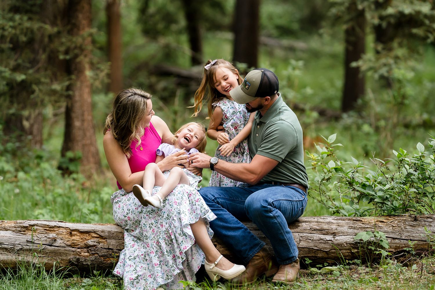 Happy family of four laughing together on a log in a lush green forest during an outdoor photo session.