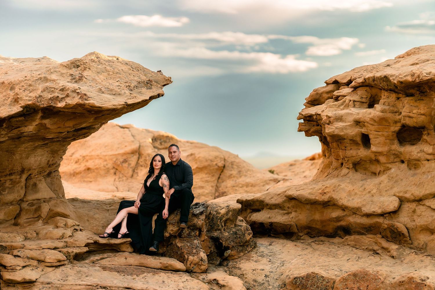 Couple in black outfits sitting together between sandstone rock formations under dramatic sky.