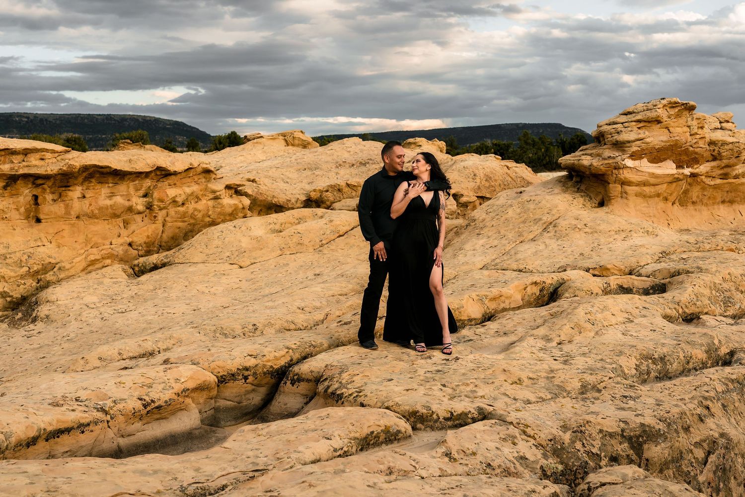 Romantic couple embracing on sun-kissed sandstone rocks with warm golden light and stormy clouds overhead.