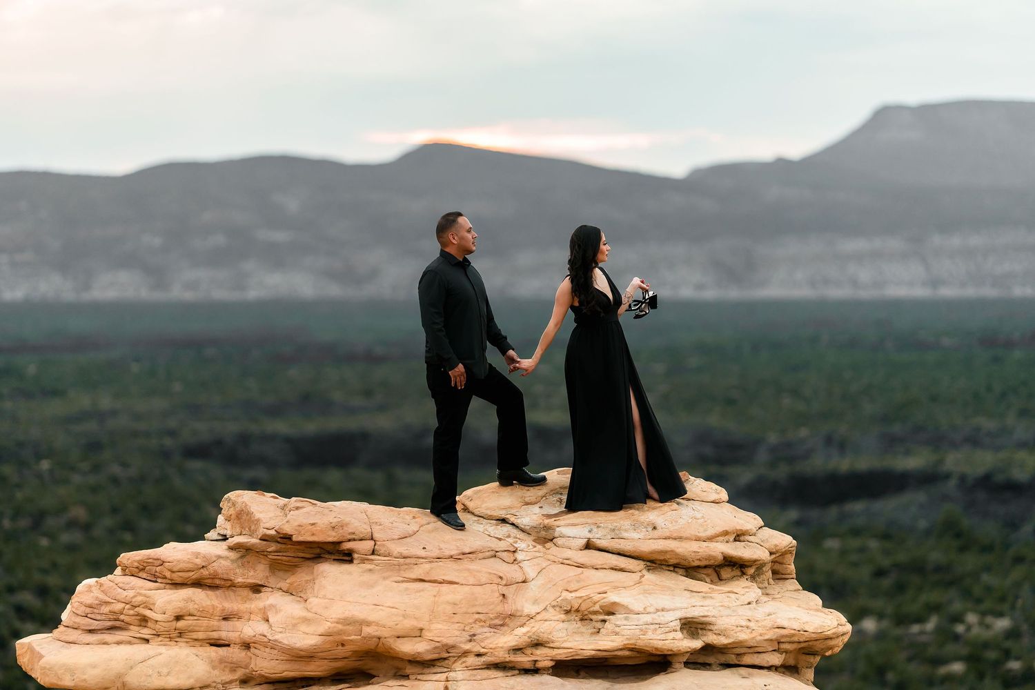Couple in matching black outfits holding hands on rocky cliff with mountain landscape backdrop.