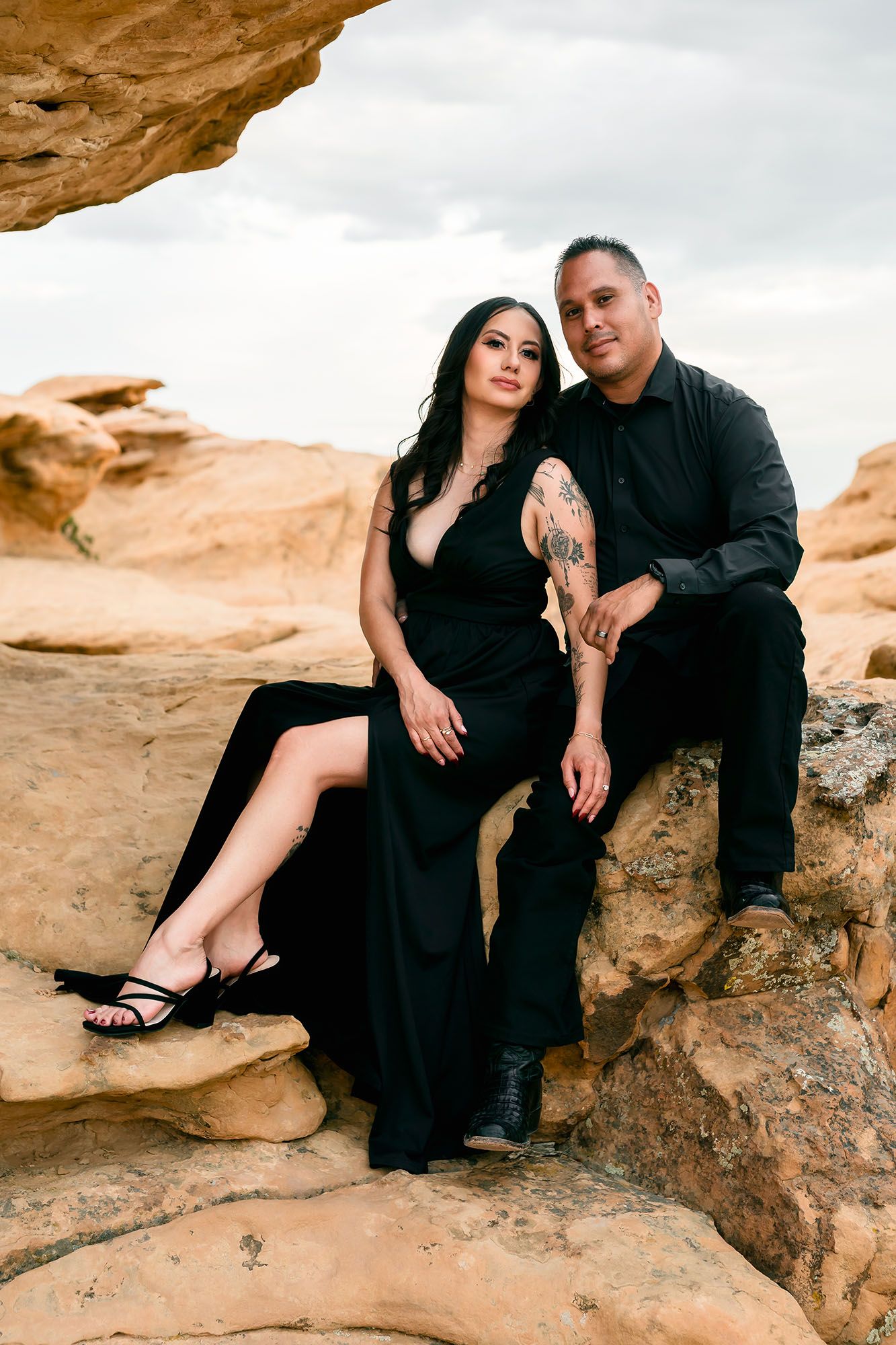 Tattooed couple in elegant black attire posing on sandstone rocks under an arch formation.