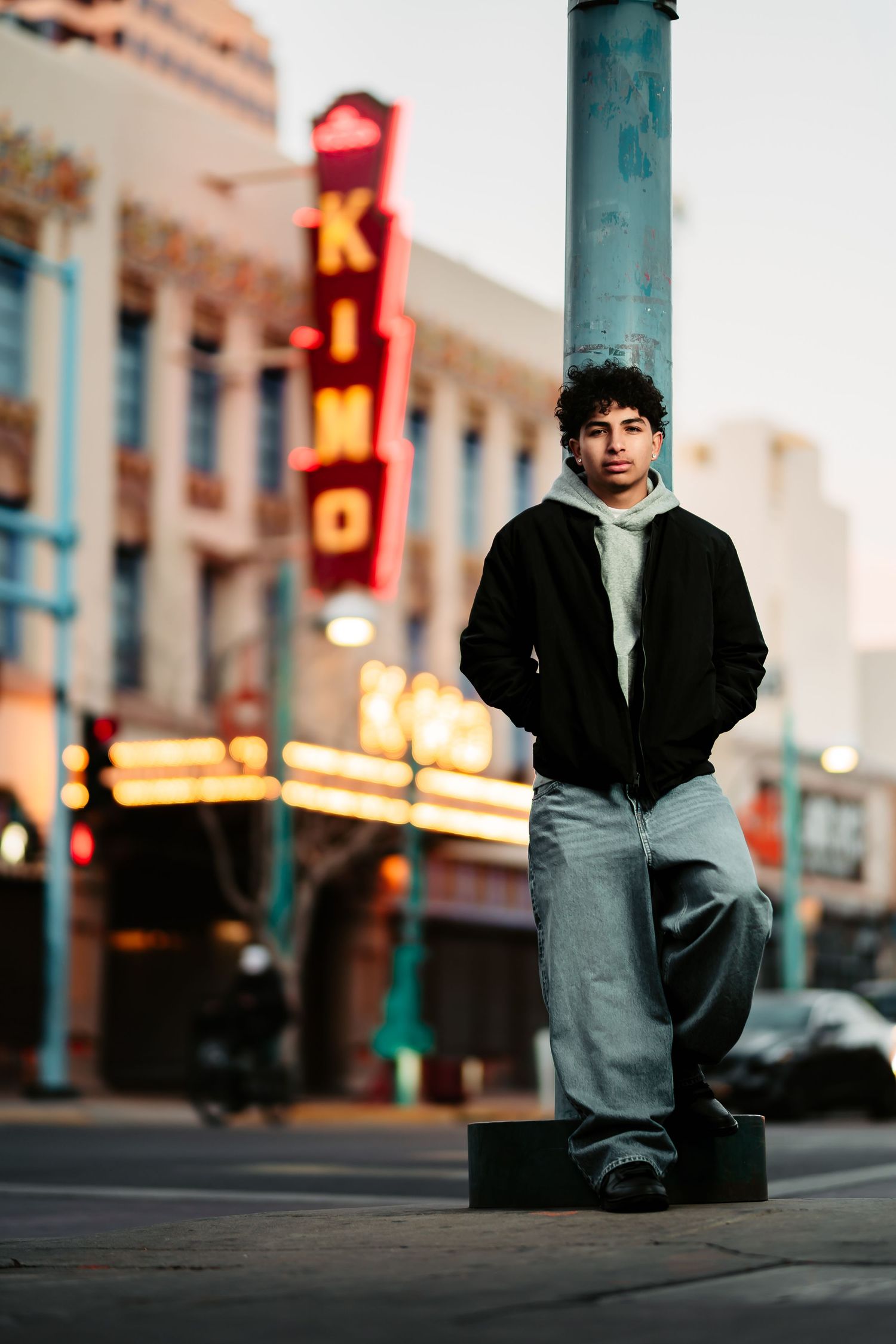 A street scene at dusk with illuminated neon signs and a figure standing on a pole base in an urban setting.