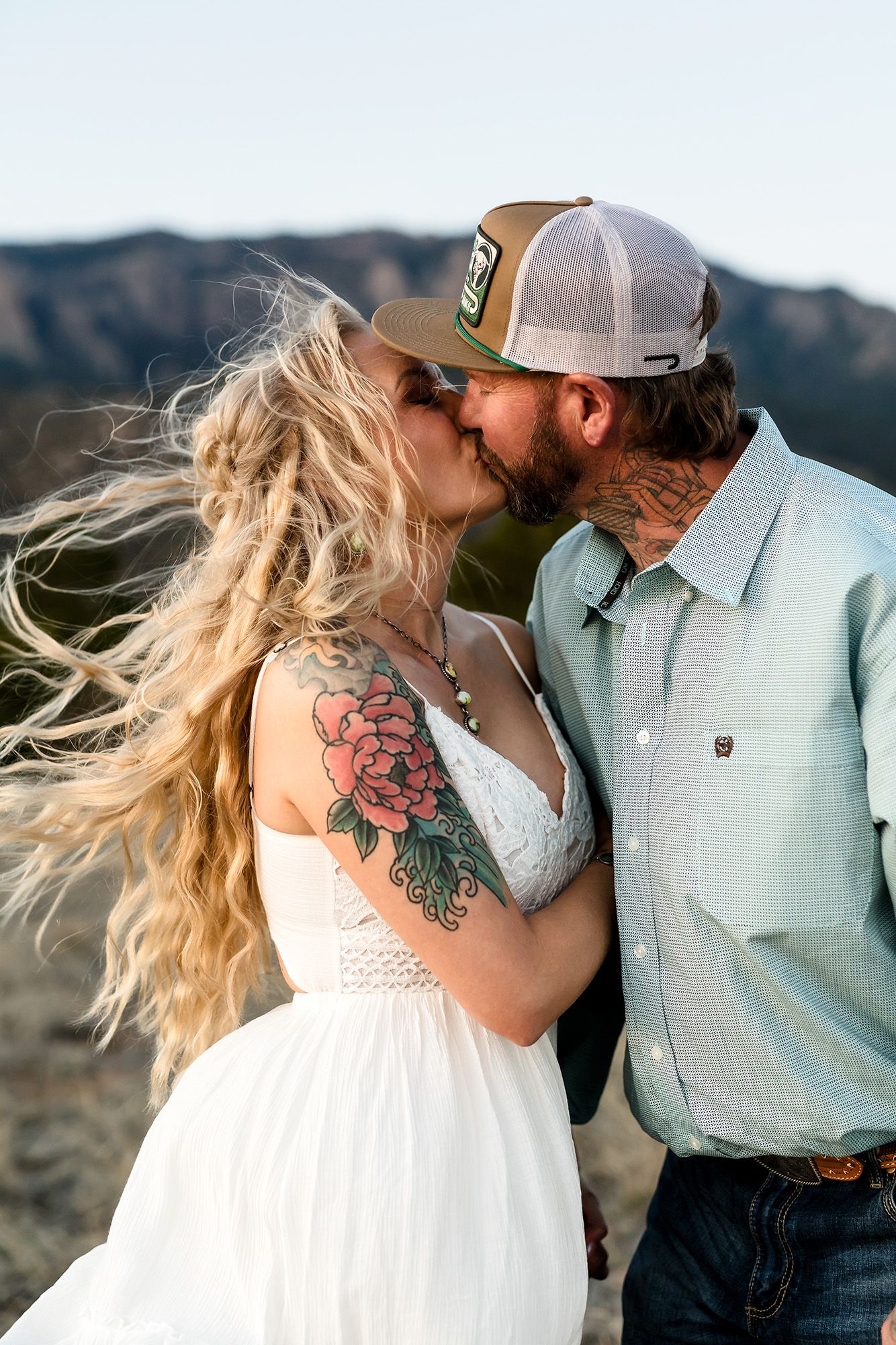 Tattooed couple sharing a romantic kiss outdoors with mountain backdrop, woman in white dress with floral tattoo, man in cap.