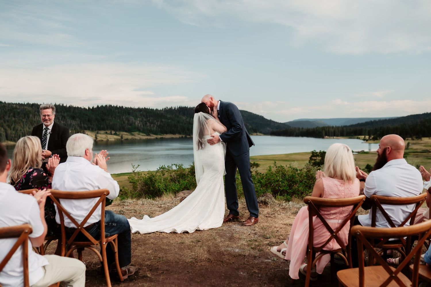 Wedding guests seated in rustic wooden chairs overlooking a scenic mountain lake on a bright sunny day.