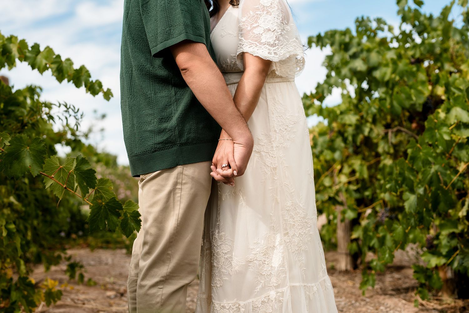 Couple holding hands in a lush vineyard, wearing a green shirt and white lace dress.