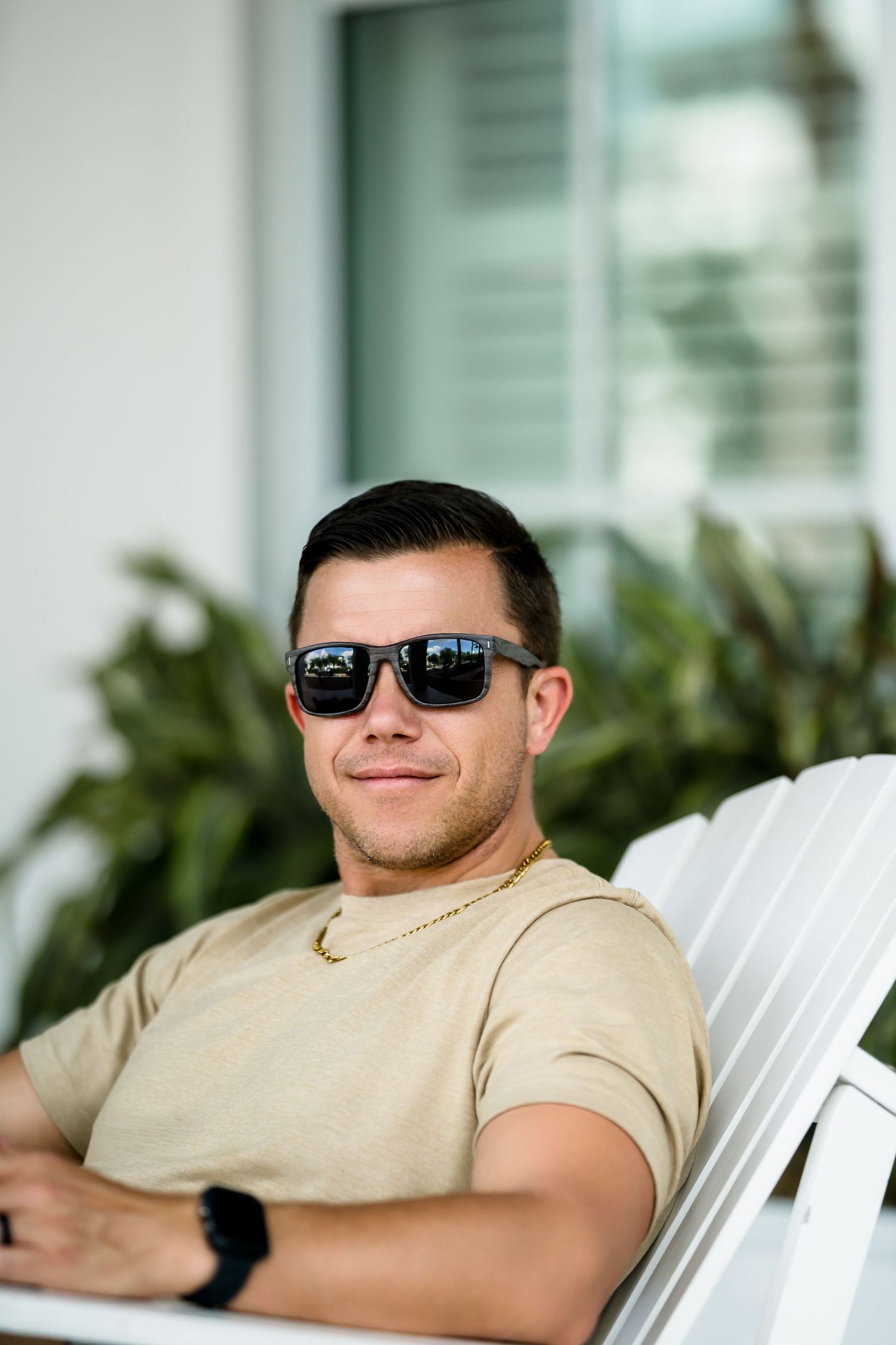 Young man in sunglasses and beige t-shirt relaxing on white chair outdoors near green plants.
