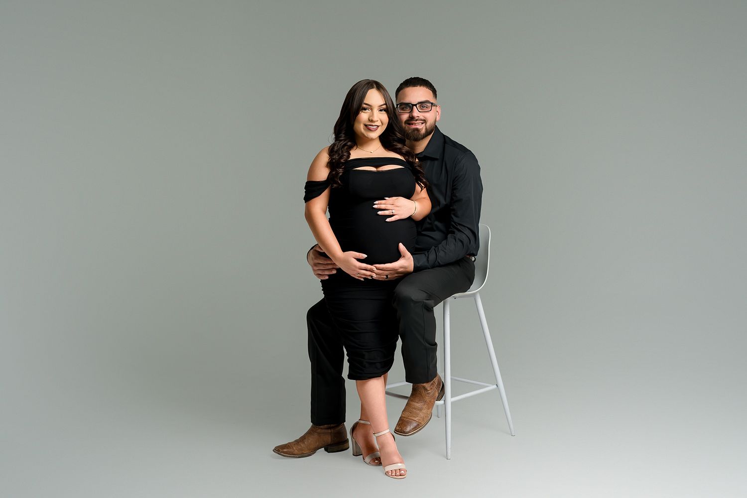 Pregnant couple posing together in black outfits on a white stool against a gray studio background.
