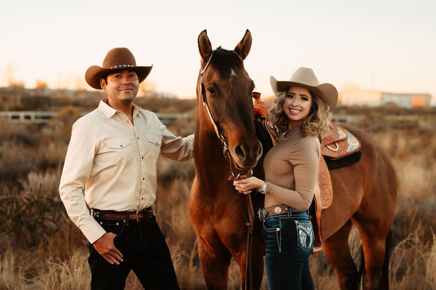 A couple in western attire shares a romantic moment with their horse during a sunset photoshoot in a desert landscape.