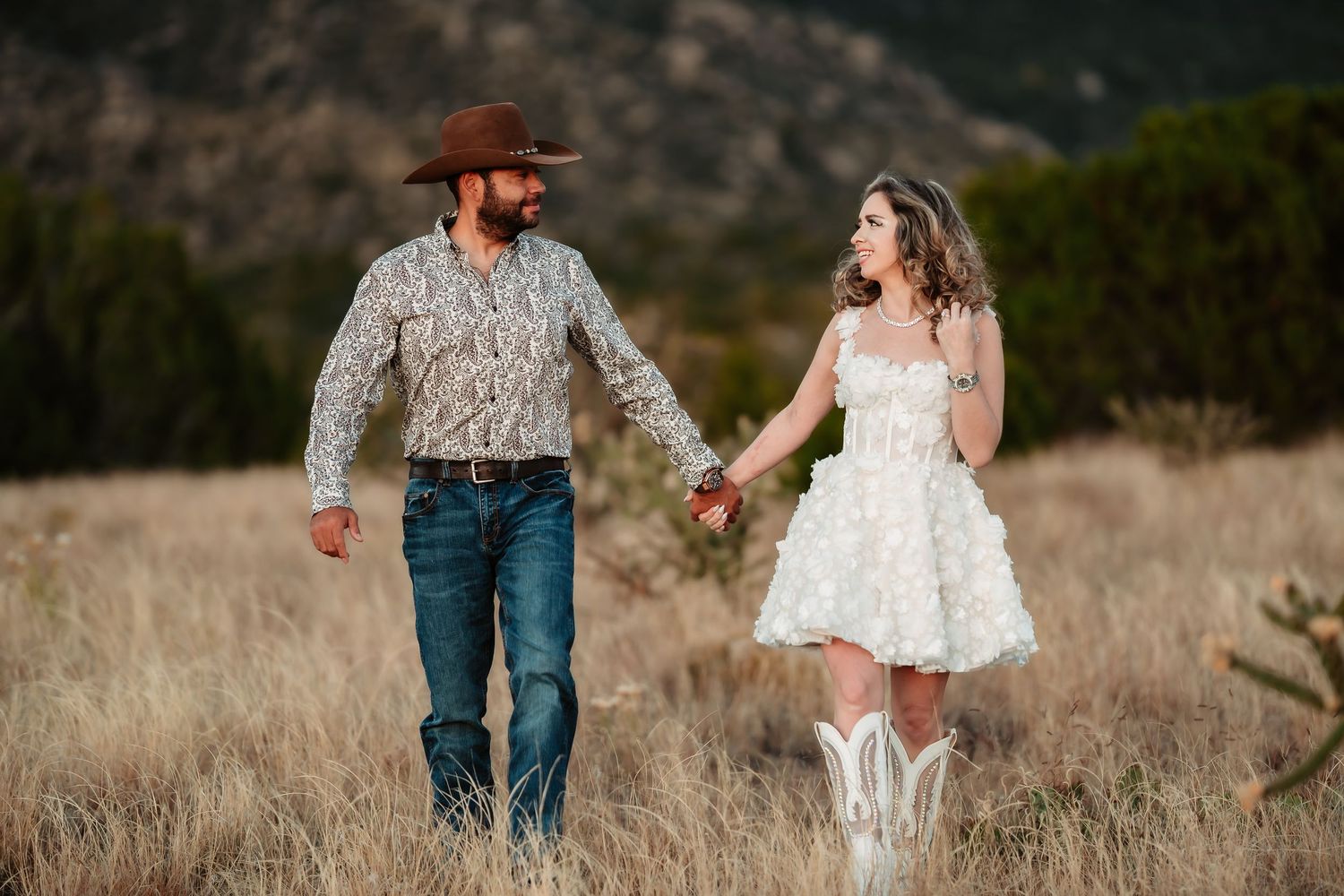 A couple in western attire walks through a golden field during sunset in the mountains.
