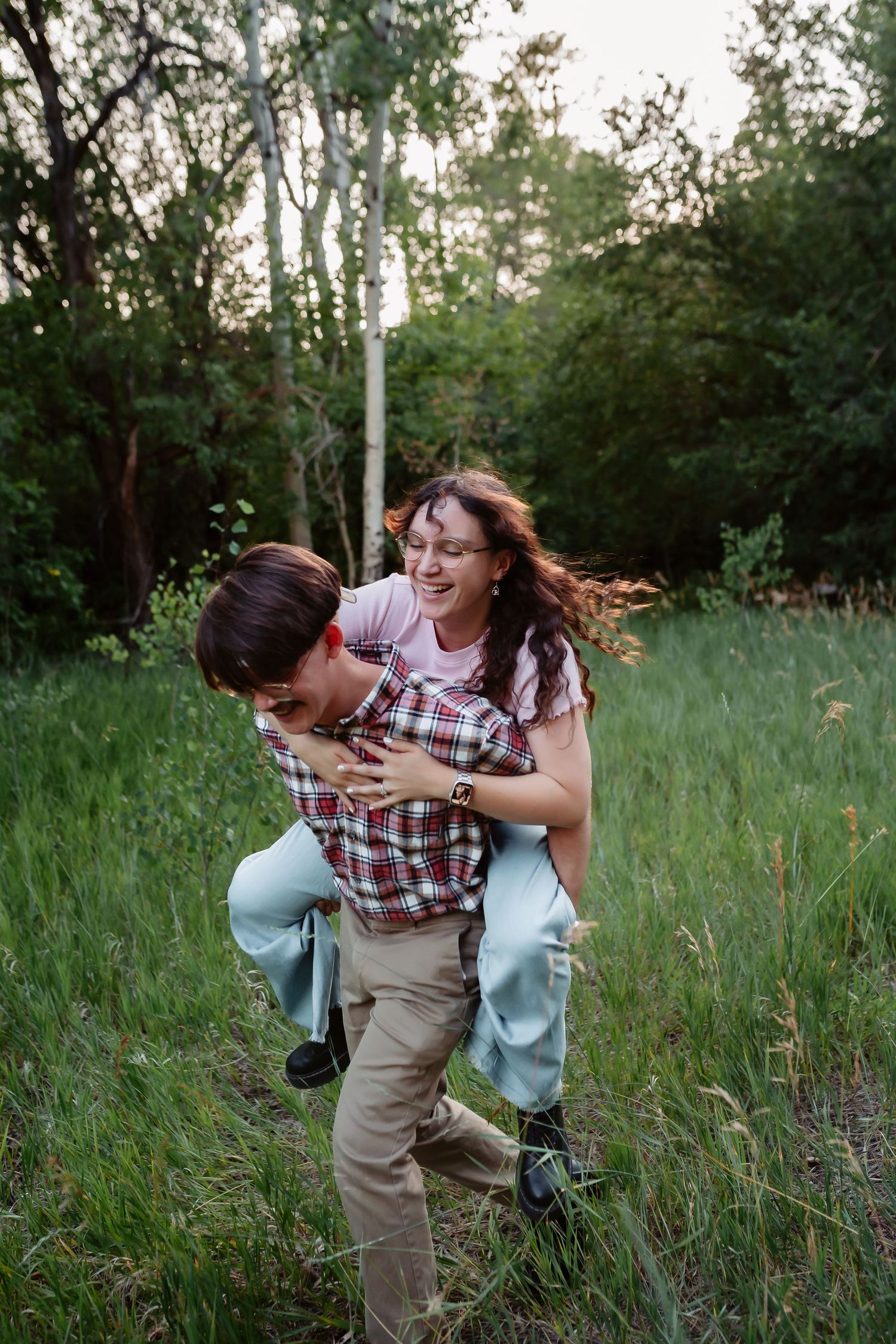 A playful piggyback ride moment captured in a lush green meadow surrounded by trees at sunset.