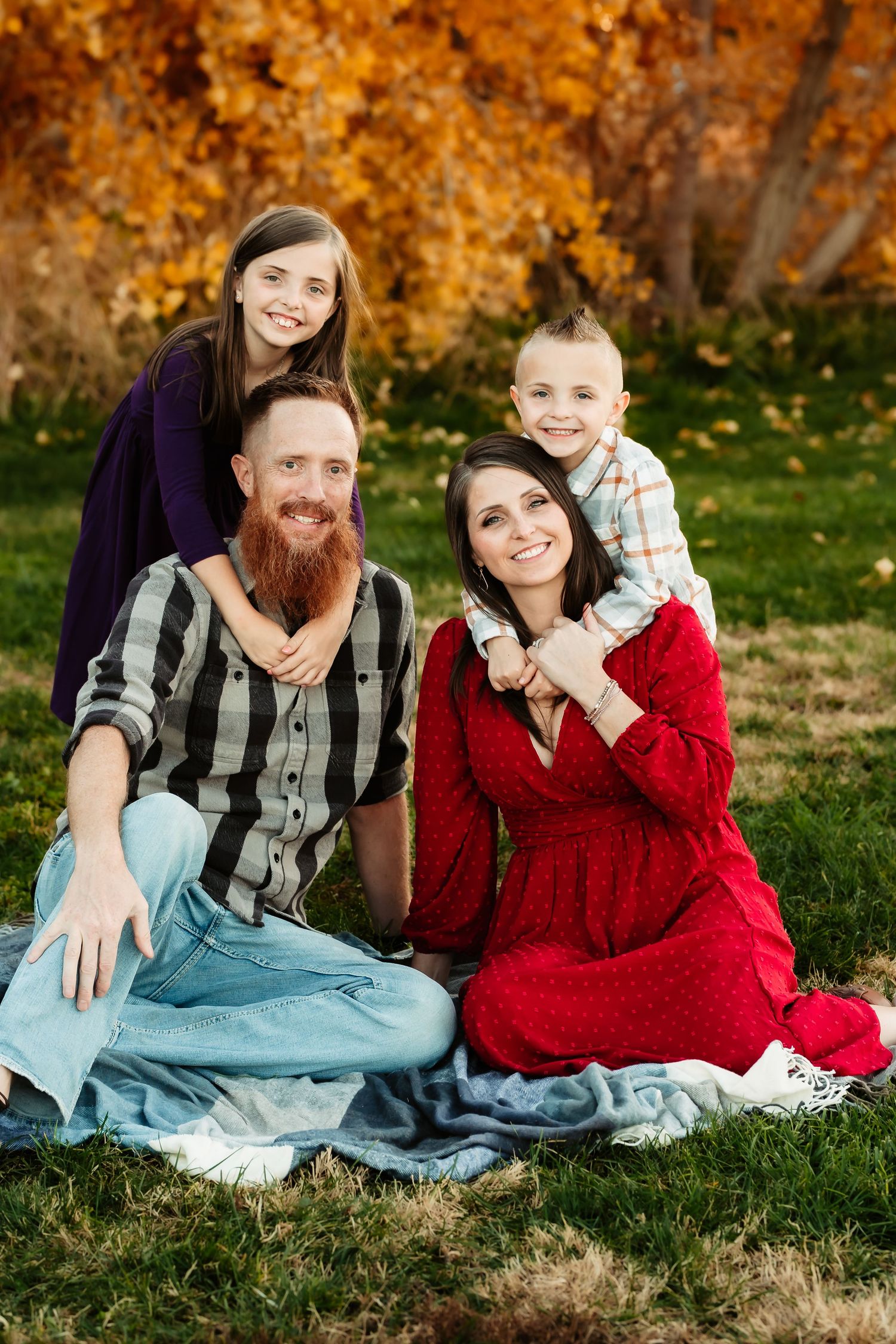 Family sitting together on grass during autumn photoshoot with vibrant fall foliage in background.
