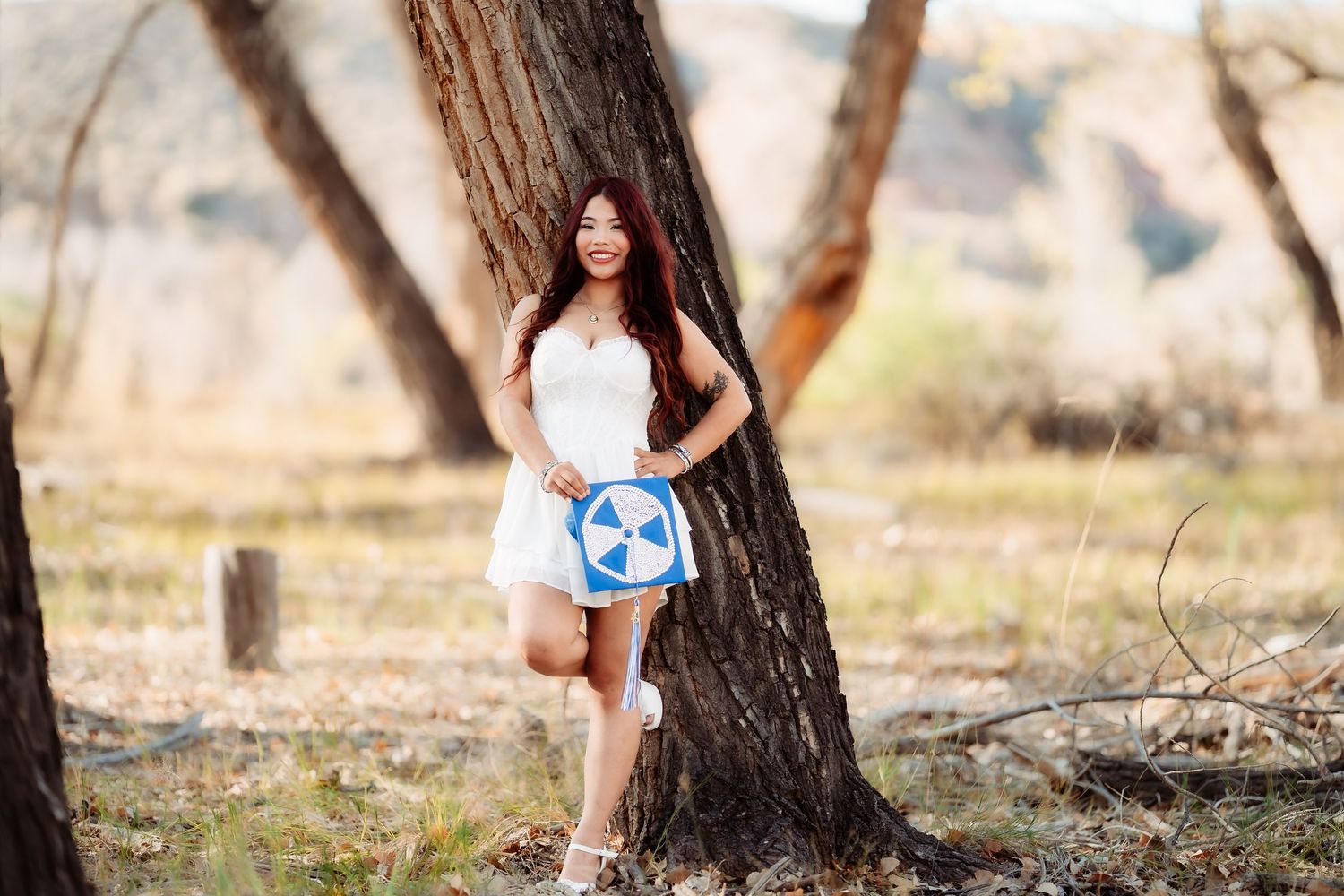 Autumn outdoor photo with person in white outfit posing with blue sign against tree.