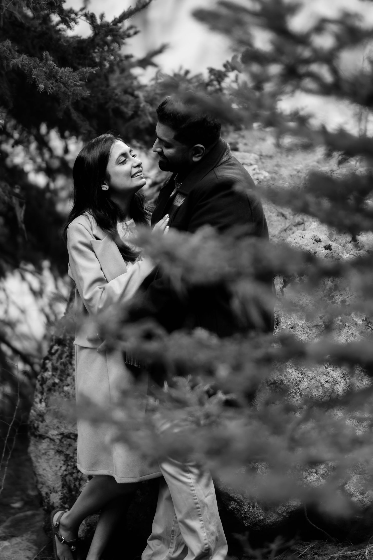 An intimate black and white portrait of a couple sharing a romantic moment among pine trees.