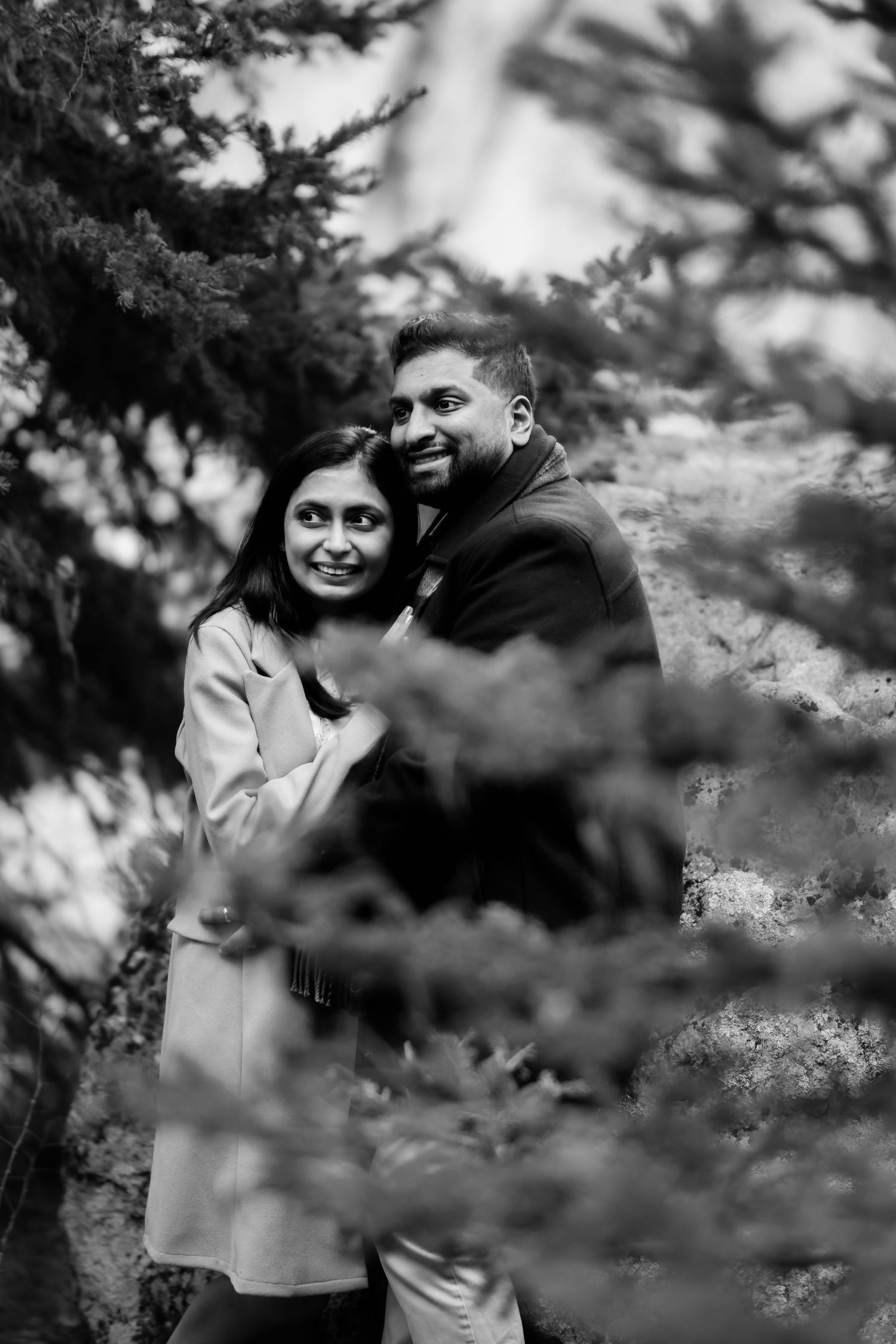 Black and white portrait of couple sharing intimate moment among evergreen trees.
