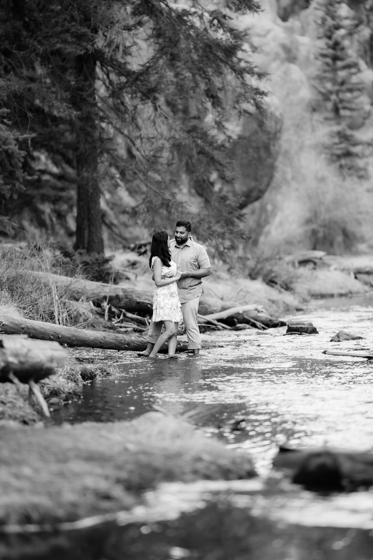 A couple standing together by a stream in a scenic forest landscape.