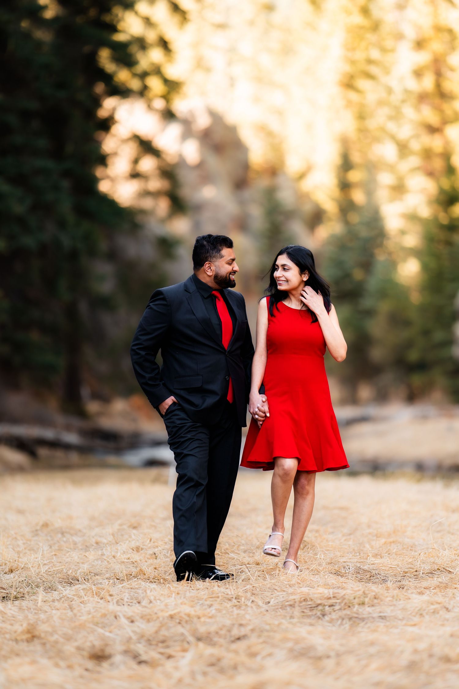 A couple in coordinating red outfits share a romantic moment while walking in a sunlit forest setting.