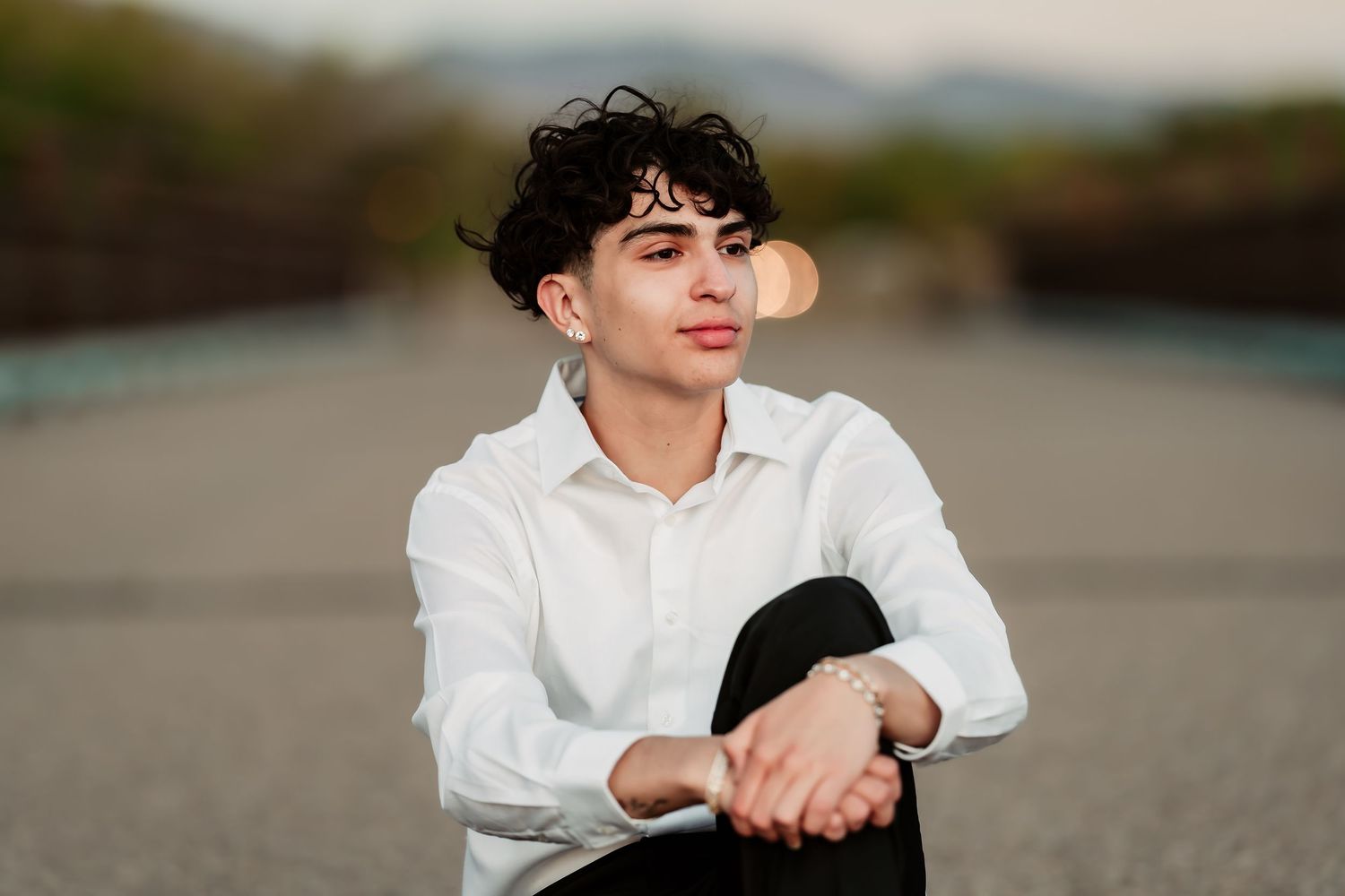 Person in a white button-down shirt sits casually on a paved road with a blurred background.