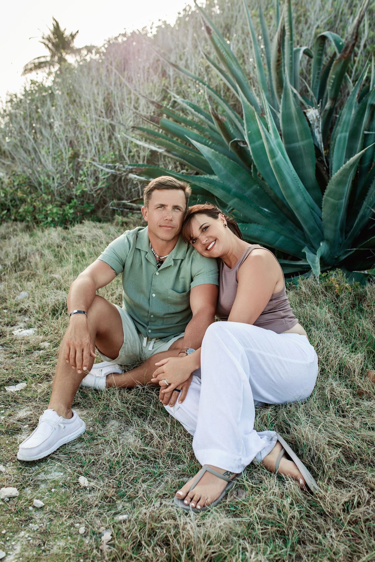 A couple sits together on grassy ground with large agave plants in the background during a romantic outdoor photoshoot.