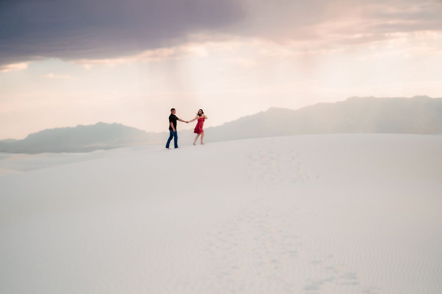 Two silhouetted figures walk across a vast white salt flat under a dramatic cloudy sky at sunset.