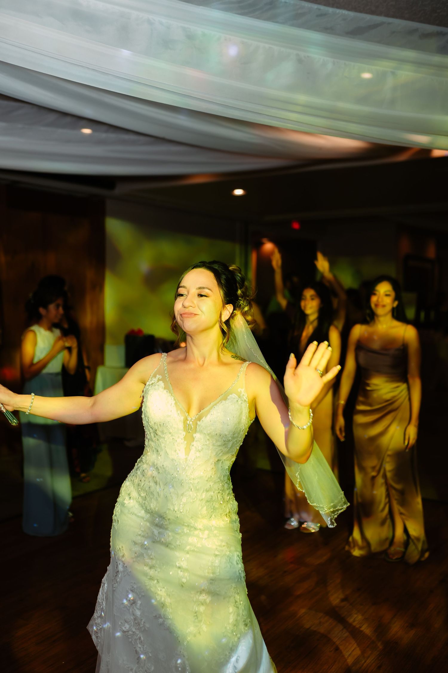 A bride in a sparkly white dress dances joyfully at her wedding reception on a wooden dance floor.
