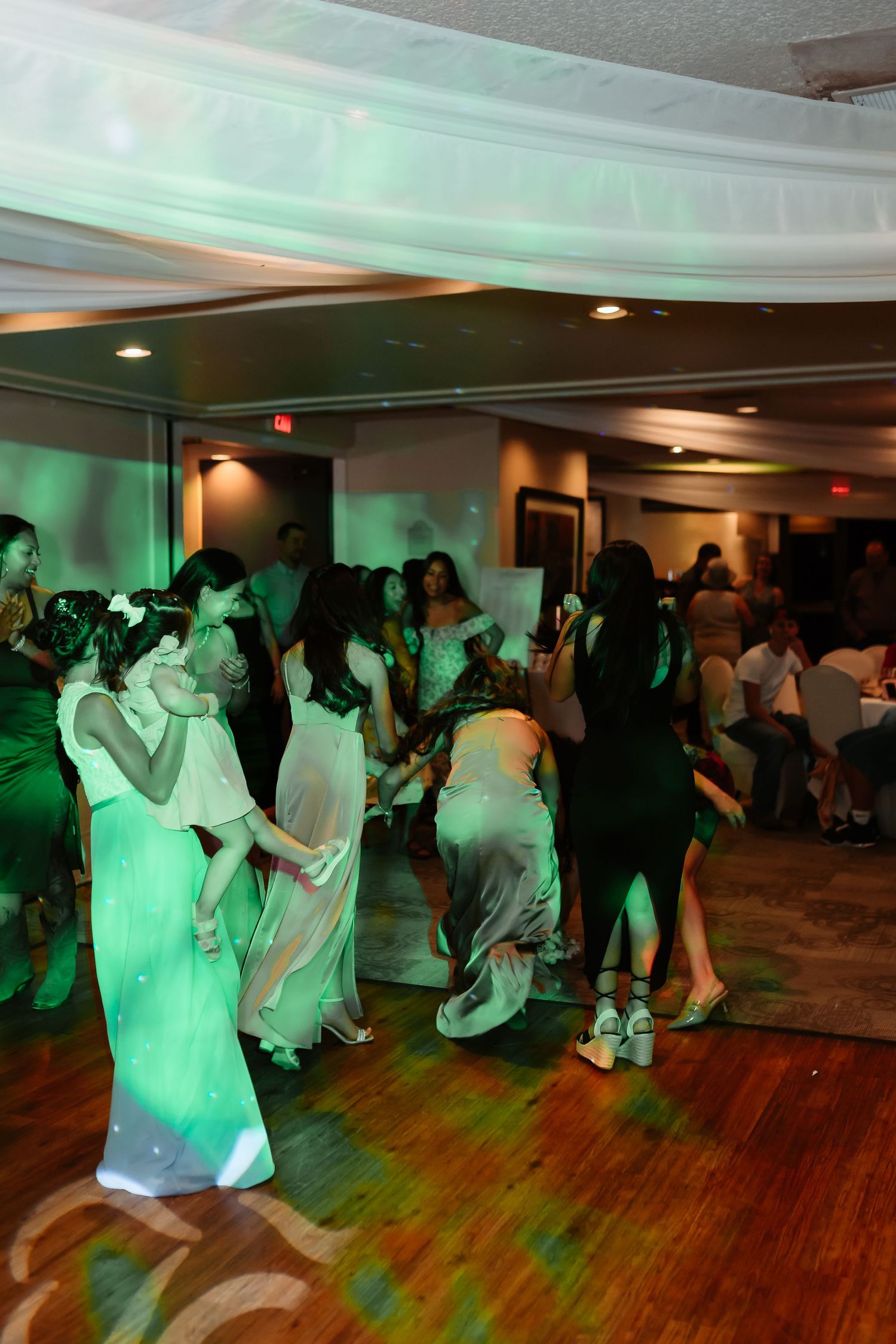 People dancing together on a wooden dance floor illuminated by green lighting at an indoor event.