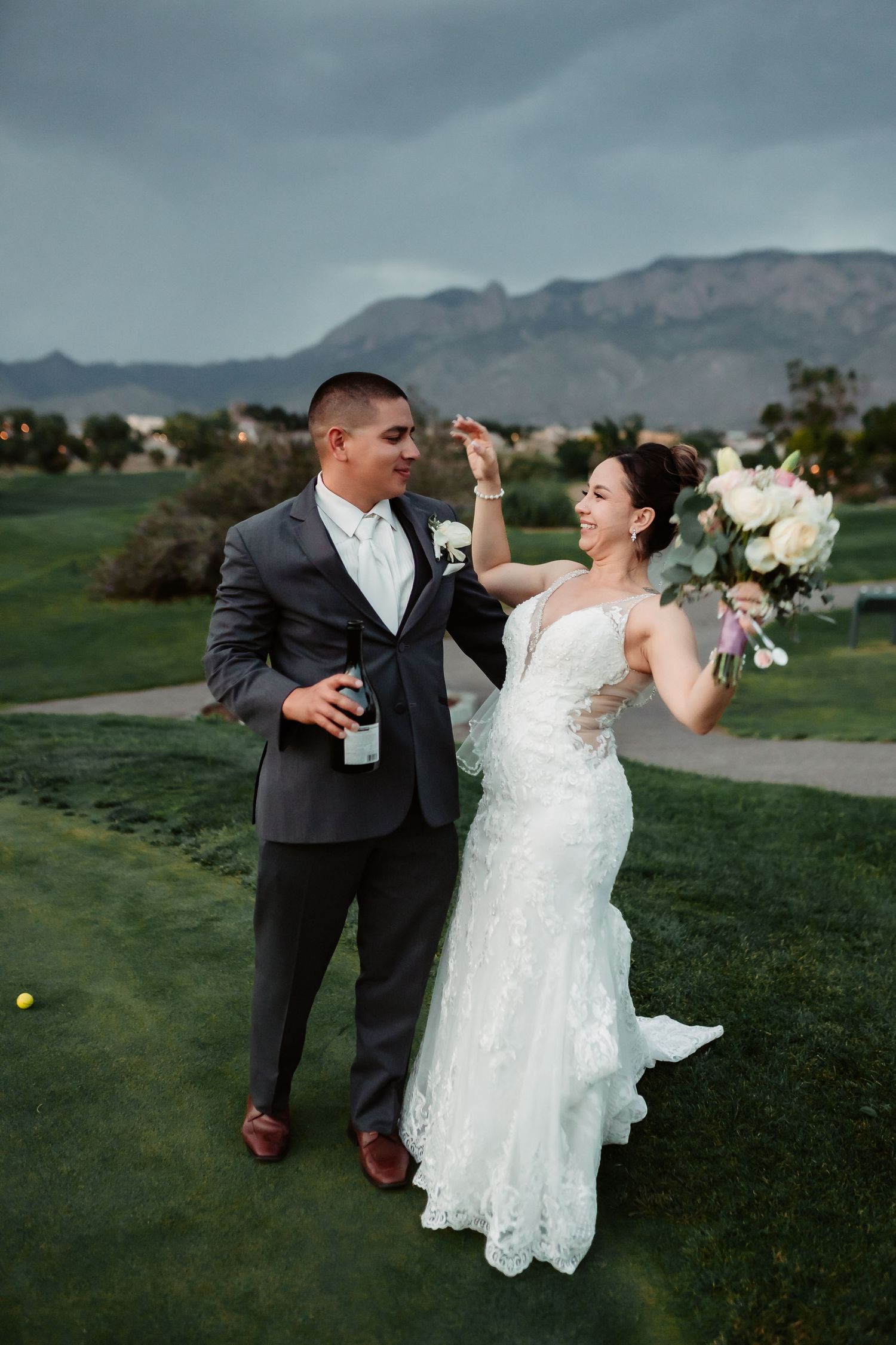 A joyful couple in wedding attire dances playfully on a lush green lawn with mountains in the background.