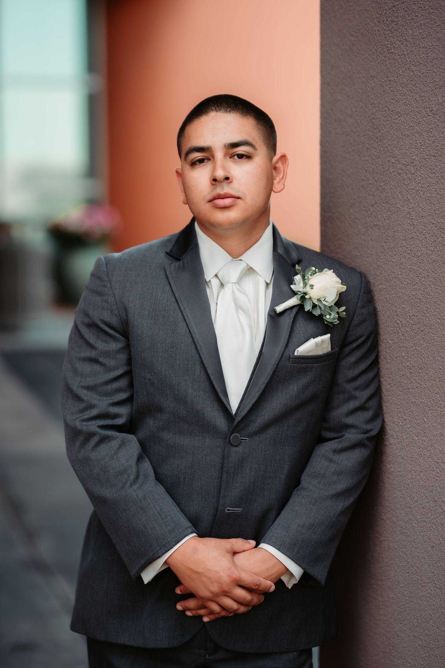 A groom in a charcoal grey suit with white boutonniere stands posed against a pink and grey wall.