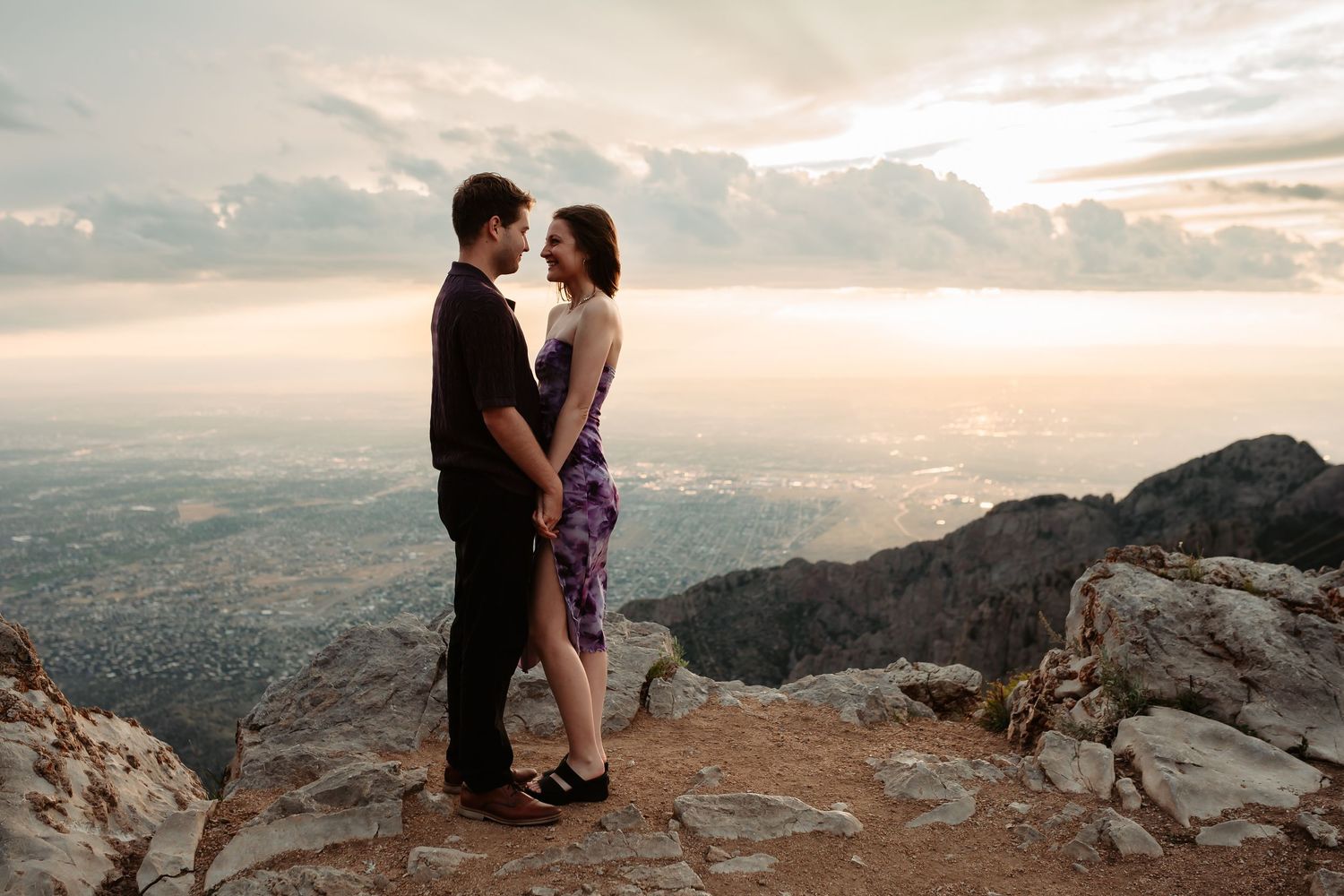 Silhouetted couple embraces at sunset on a rocky mountain overlook with dramatic cloudy skies.