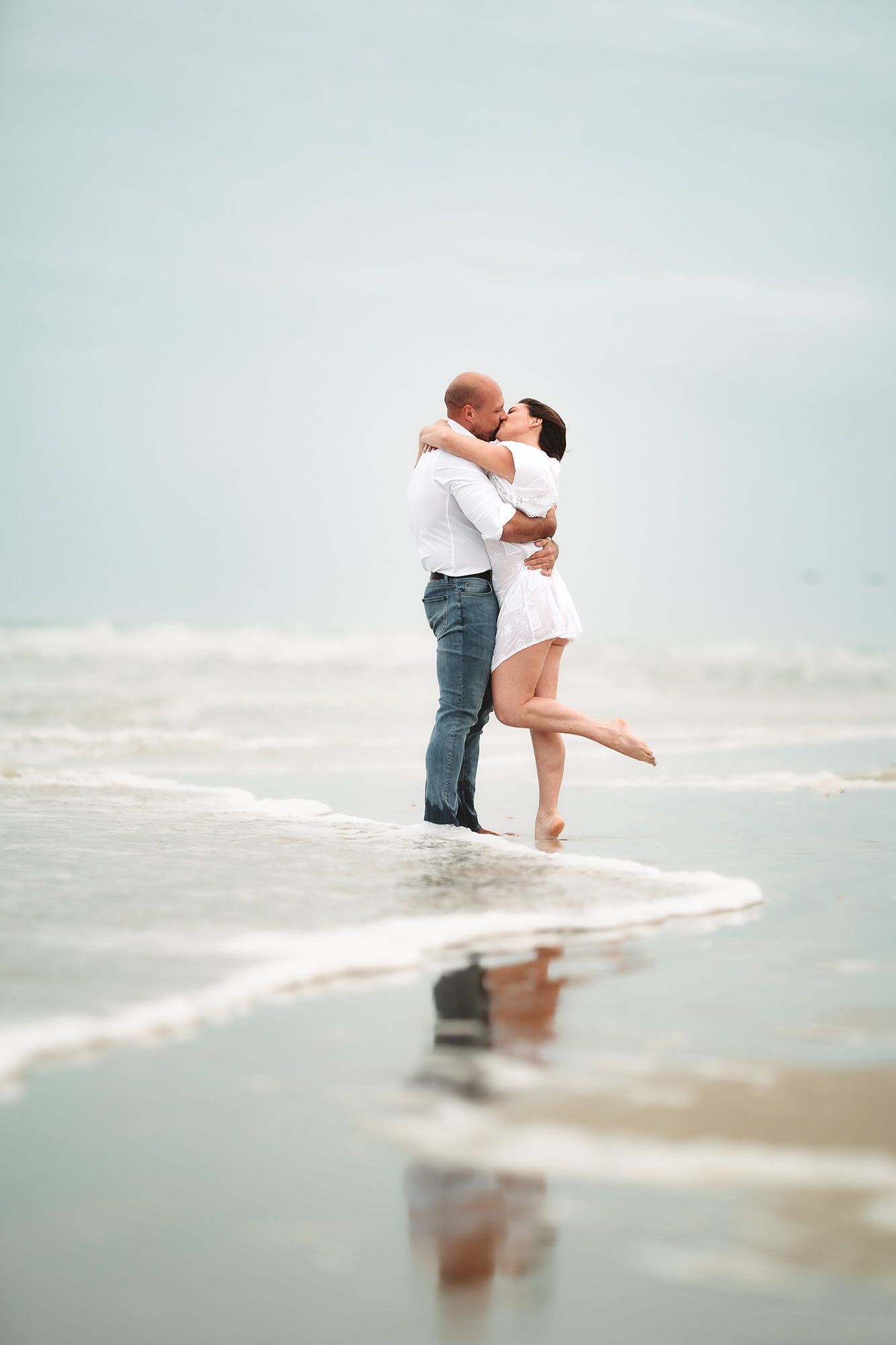 Romantic couple kissing on a beach shoreline, waves gently surrounding them as woman lifts one leg in a tender embrace.
