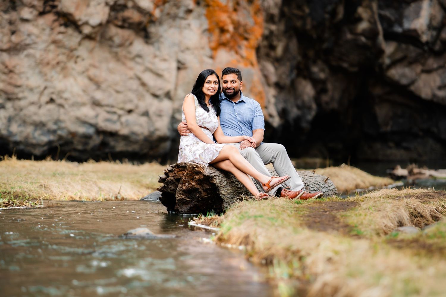 Couple sits together on rocky riverbank with cliffs and water in scenic outdoor setting.