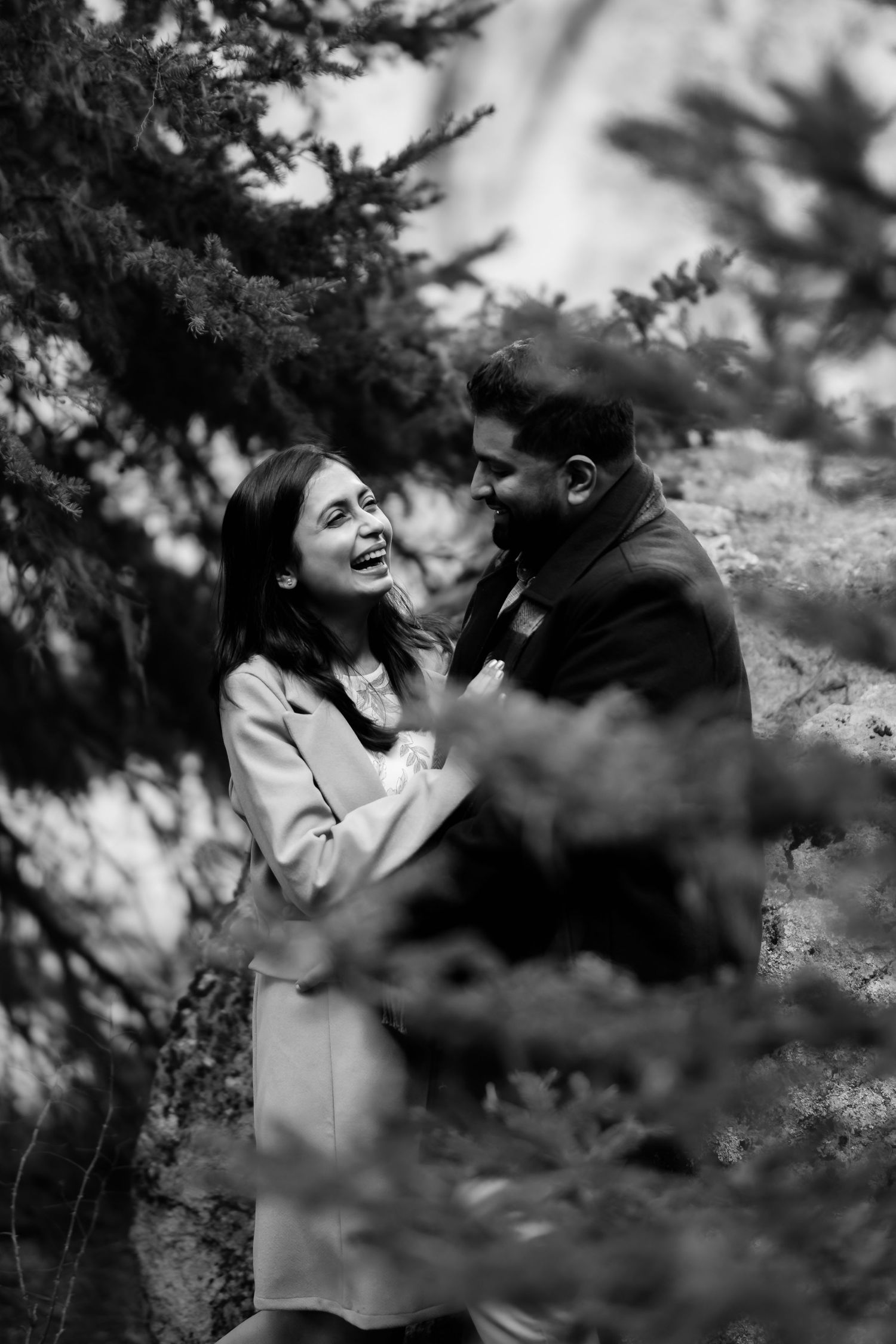 A romantic black and white photo showing a couple sharing an intimate moment among evergreen trees in nature.
