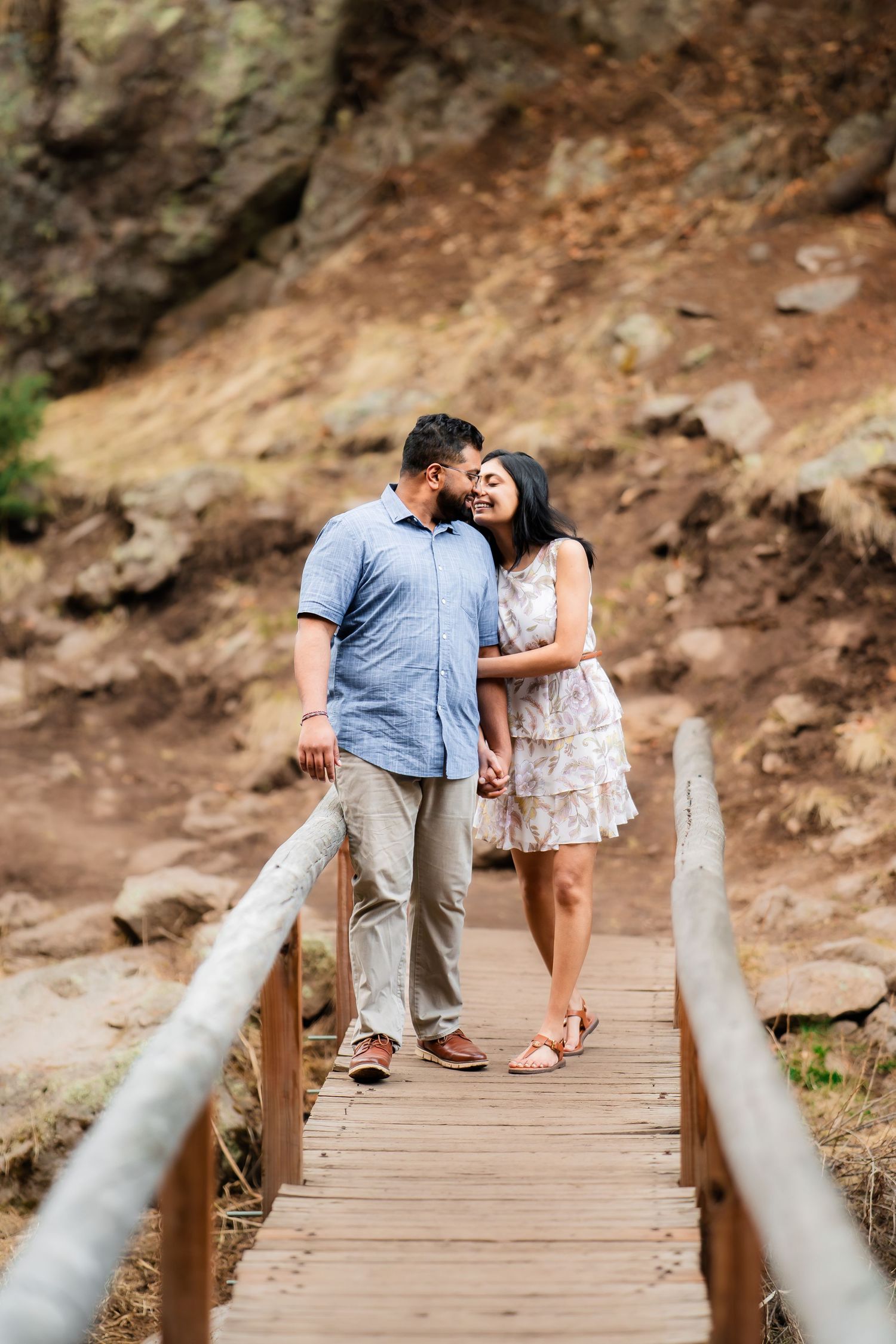 A couple stands together on a wooden walkway against a backdrop of rocky cliffs and nature.