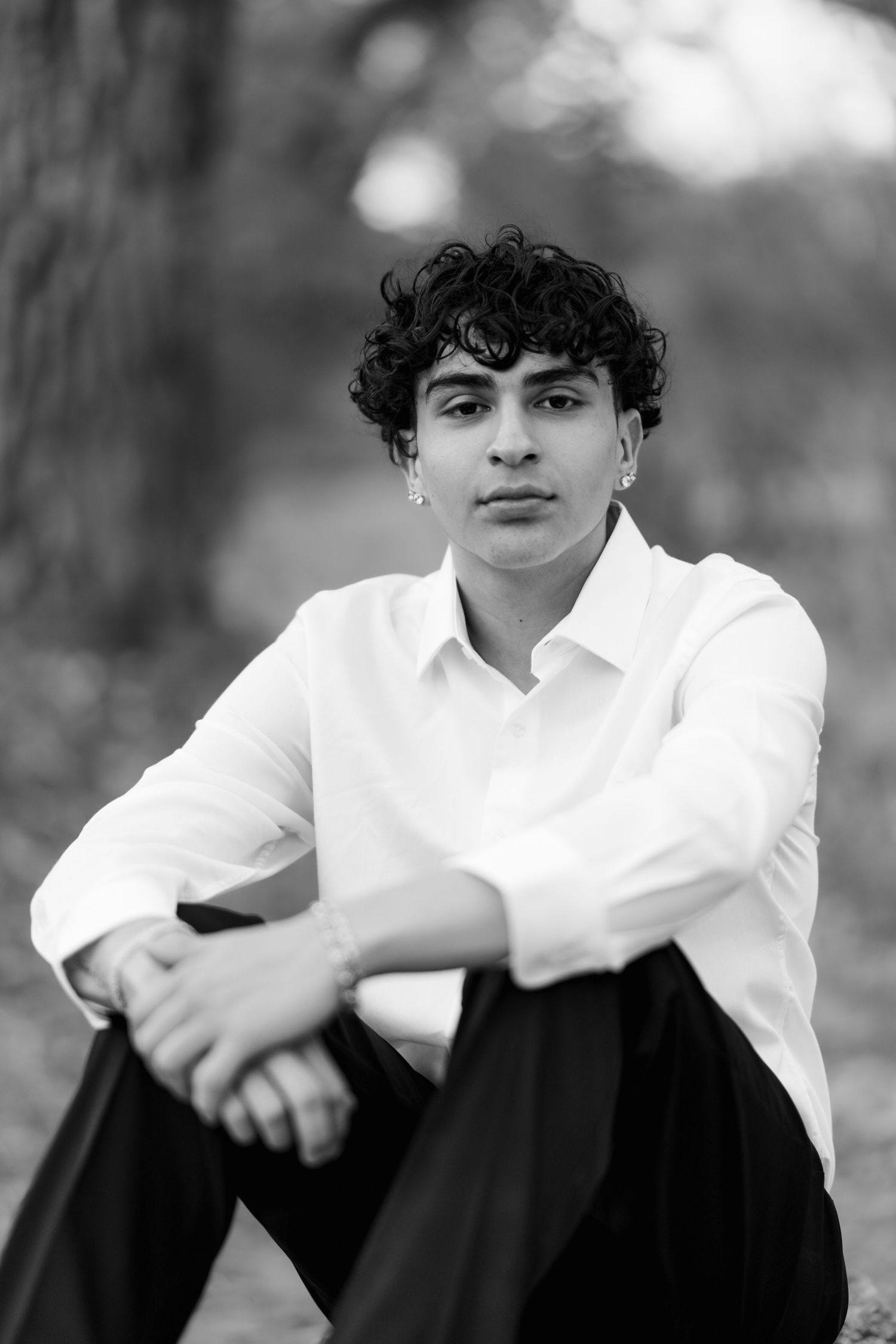 A black and white portrait of a young person in a white button-down shirt sitting outdoors with curly hair.