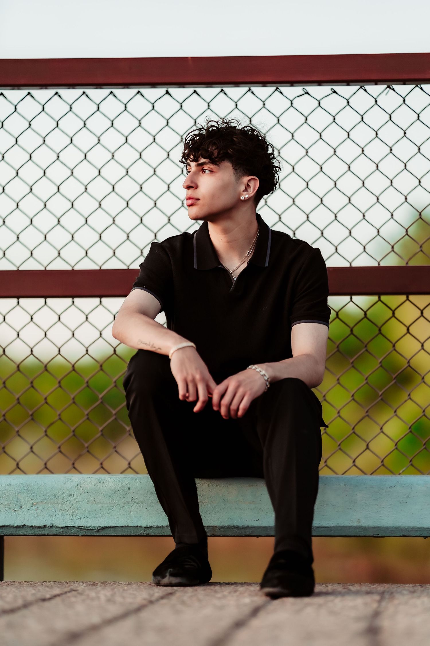 A person in a black polo shirt sits contemplatively on a bench against a chain-link fence during daytime.