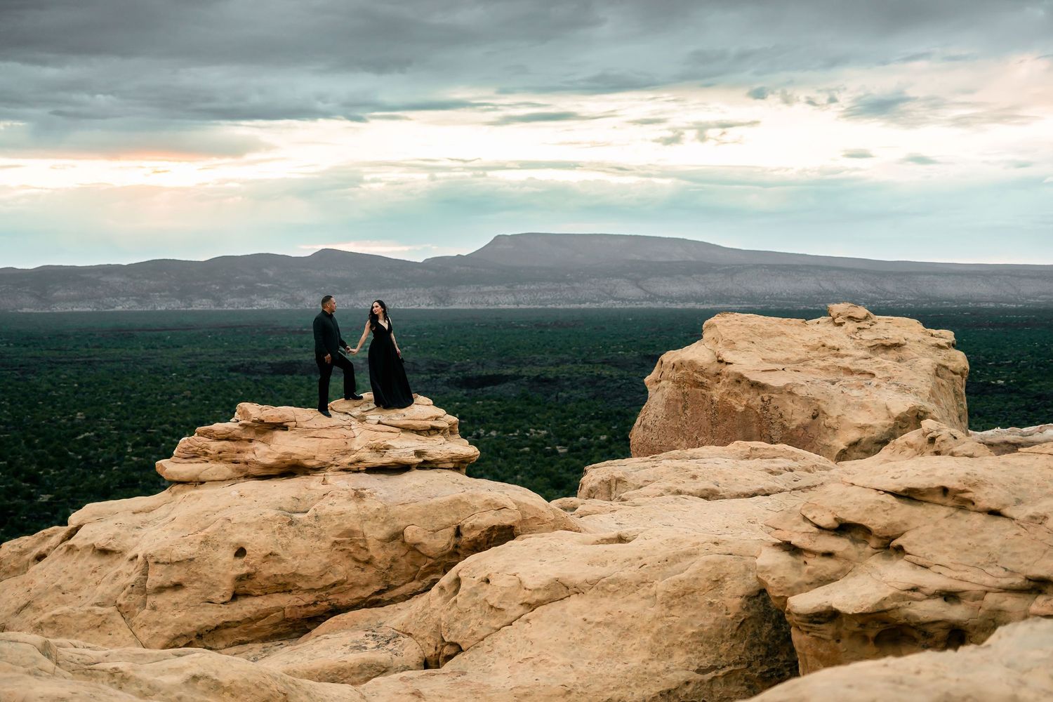 A couple stands on sandstone rocks overlooking a vast desert valley with mountains at sunset.