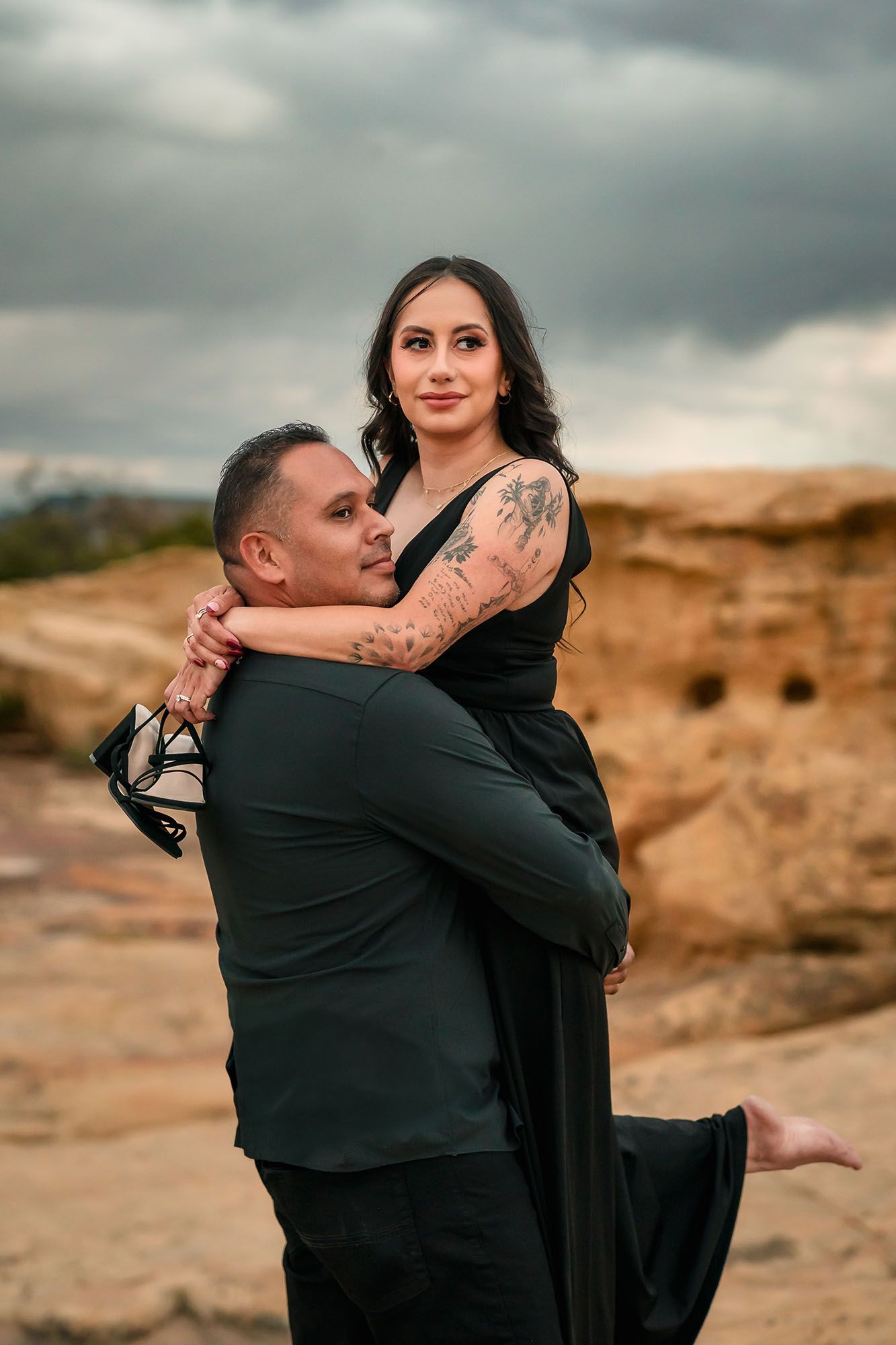 A man lifts a tattooed woman in a black dress as she wraps her arms around him on desert rocks.
