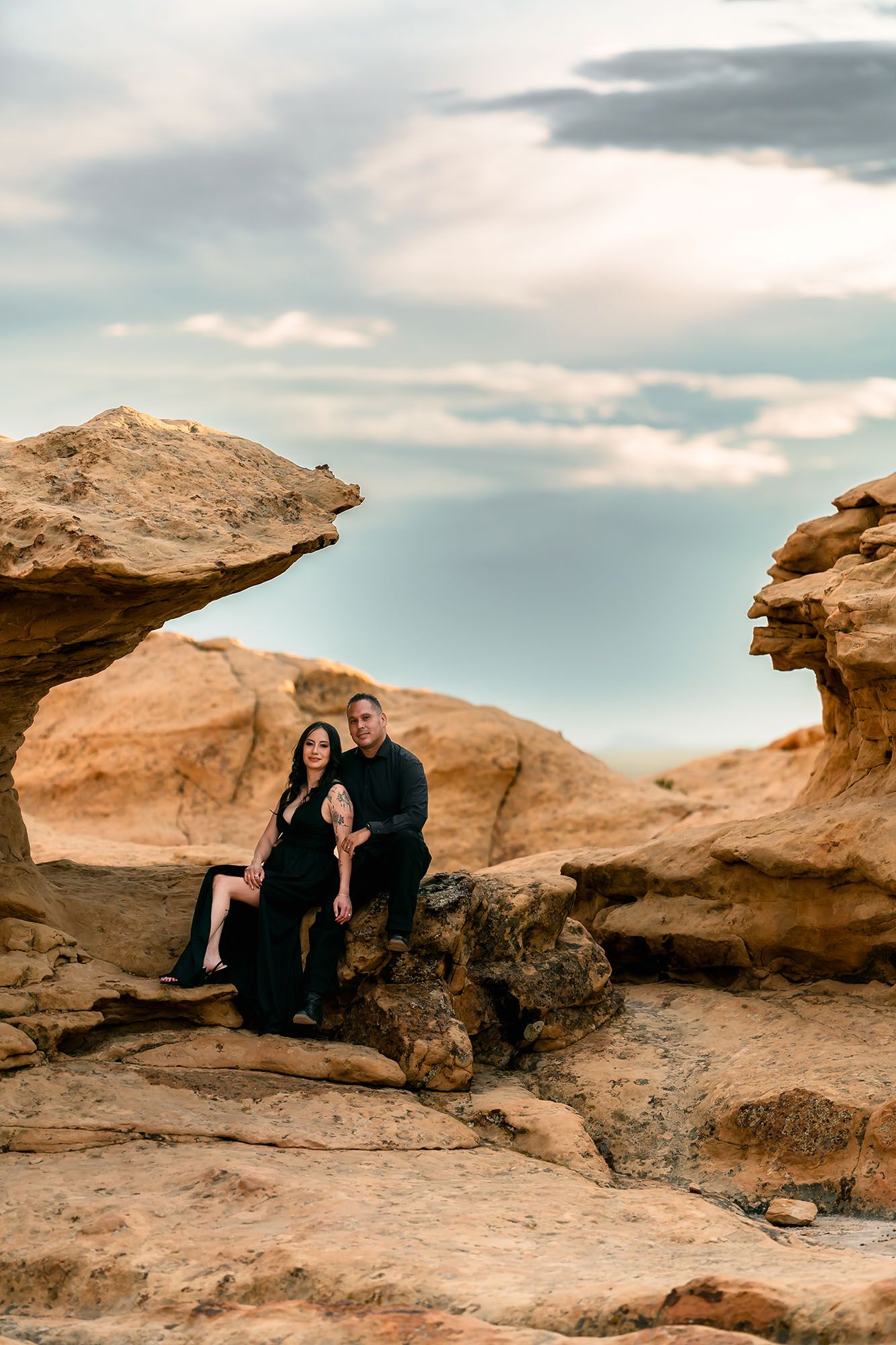 Couple in black outfits sitting among sandstone rock formations under dramatic cloudy sky.