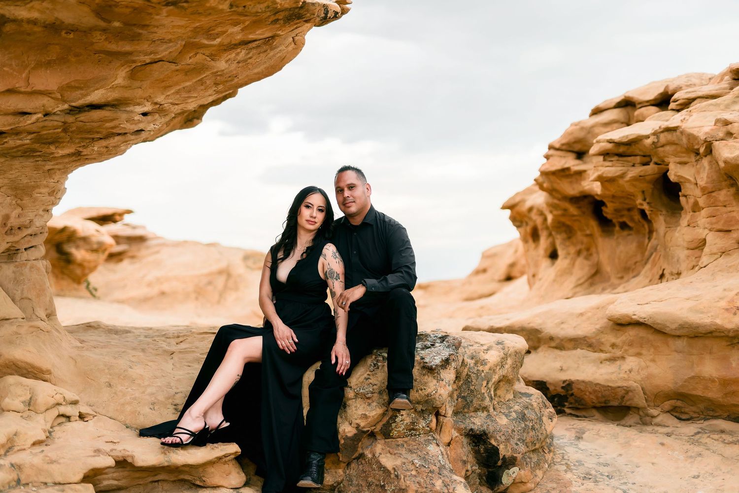 Couple in black outfits sitting together under natural rock arch in desert sandstone landscape.