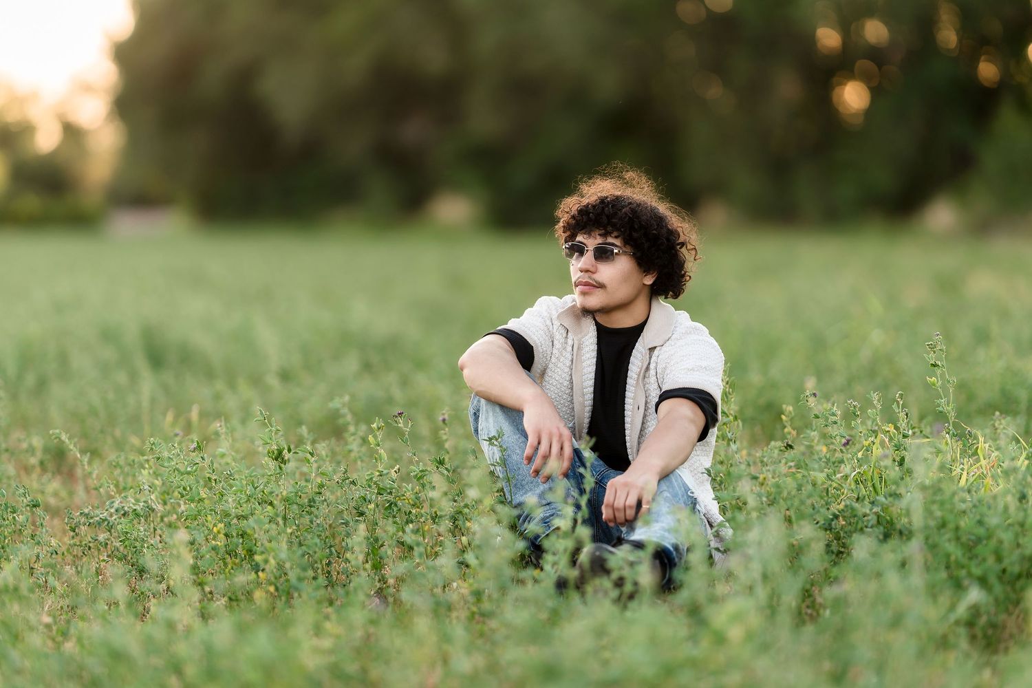 Young man with curly hair and sunglasses sitting in a green field at sunset wearing jeans and a vest.