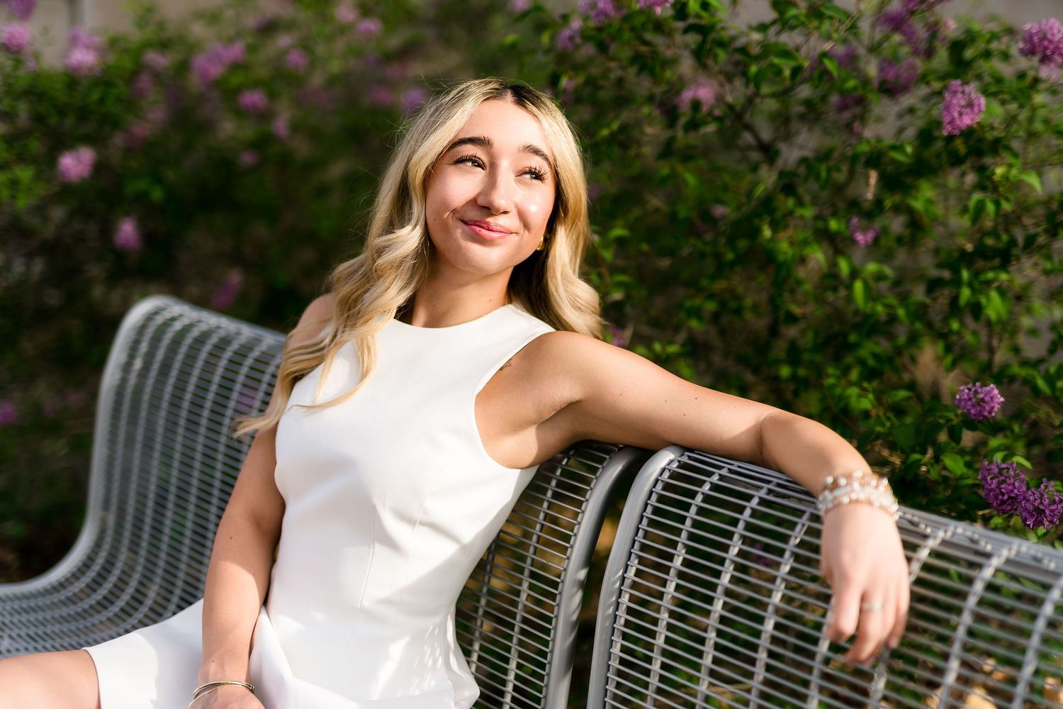 Smiling blonde woman in white sleeveless dress sits on bench with purple flowers behind her.