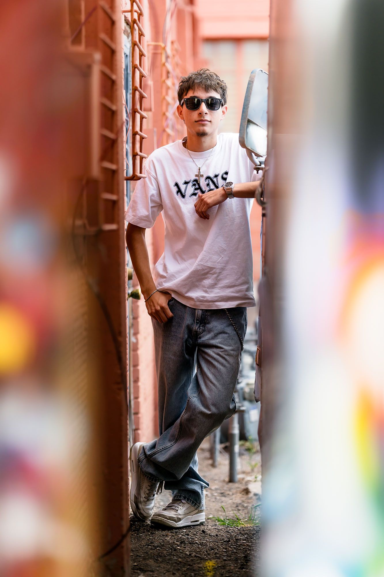 Young man in Vans t-shirt and sunglasses leaning against a wall in an urban alley setting.