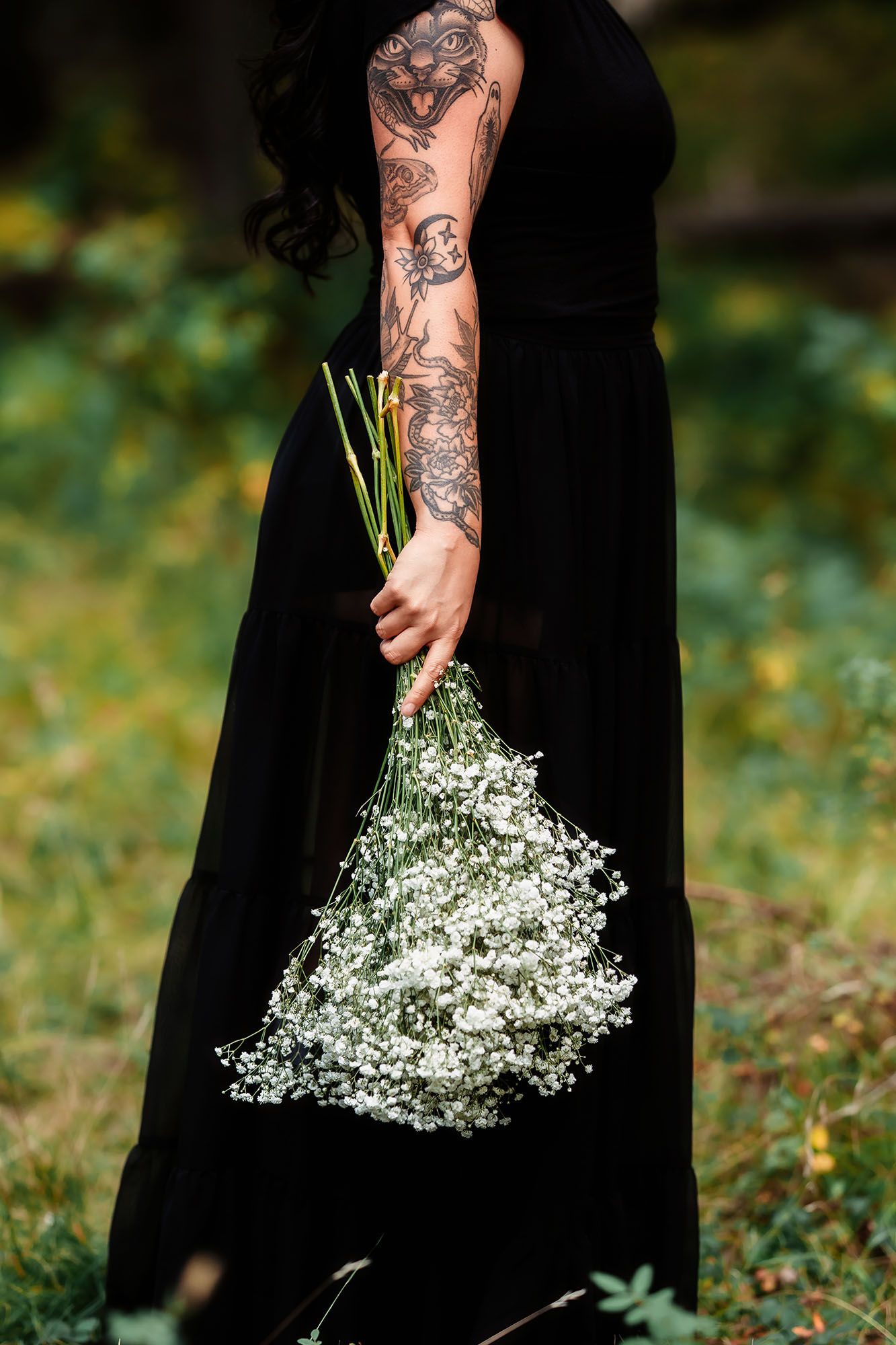 Tattooed woman in black dress holding white baby's breath flower bouquet in a lush green outdoor setting.