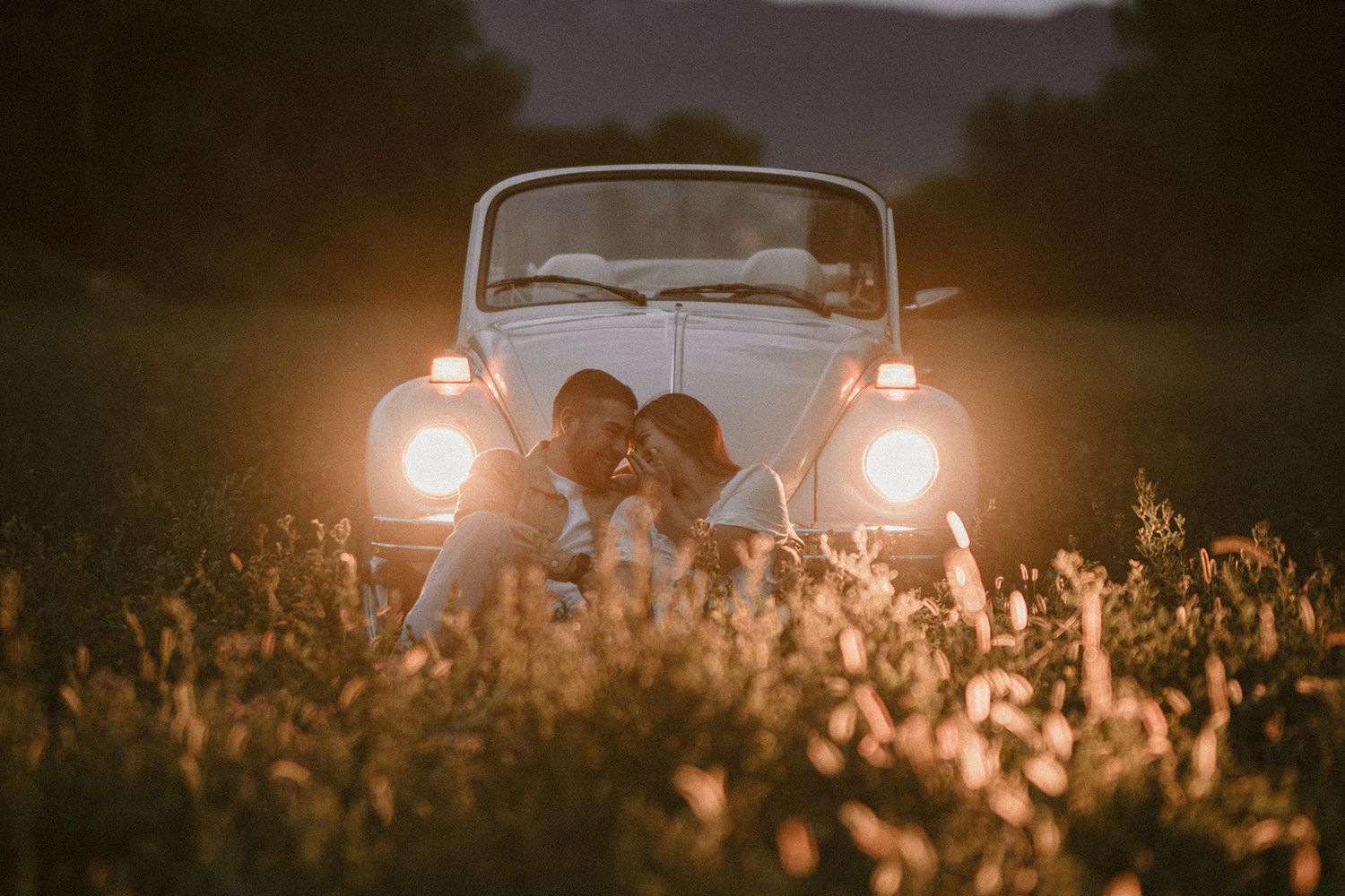 Couple sitting in a wildflower field at dusk in front of a vintage VW Beetle convertible with glowing headlights.