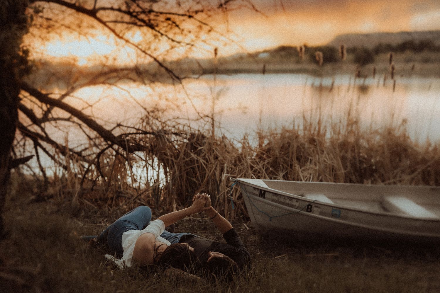 Person lying in grass by a boat near a lake at golden sunset hour.