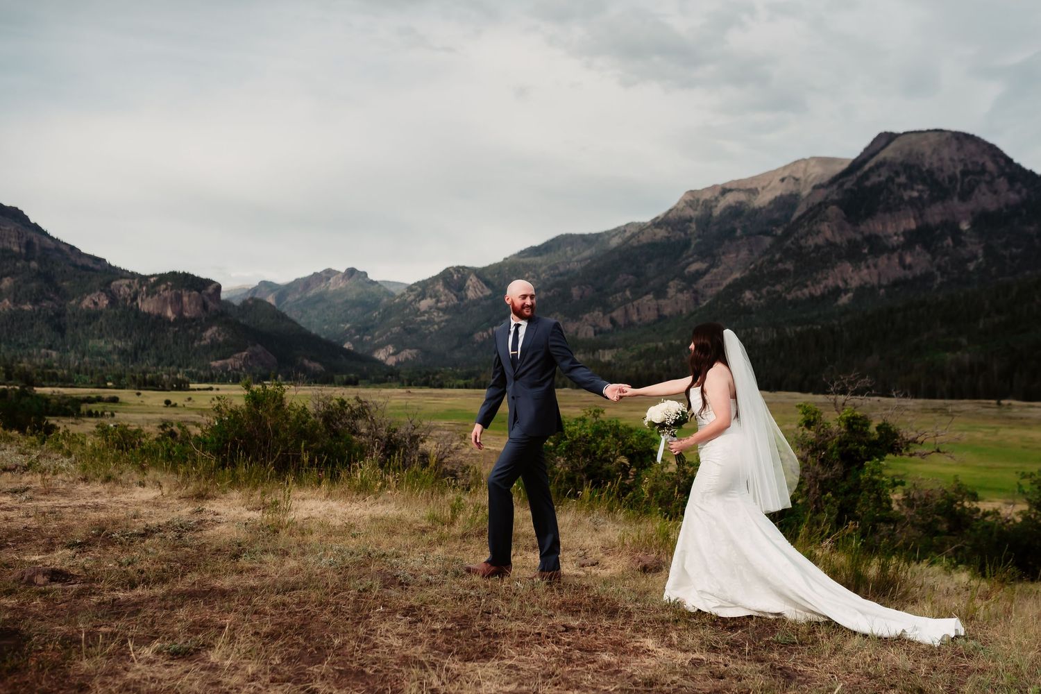 A couple in wedding attire stands in a meadow with dramatic Rocky Mountain peaks rising in the background.