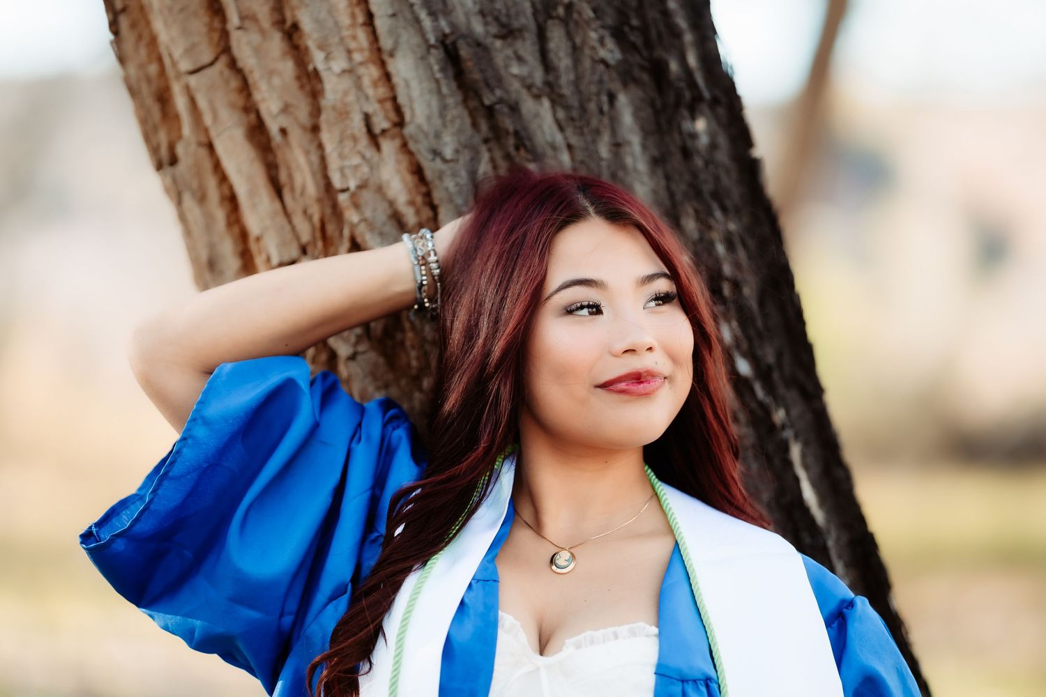 A person in blue and white attire poses near a large tree trunk in a natural outdoor setting.