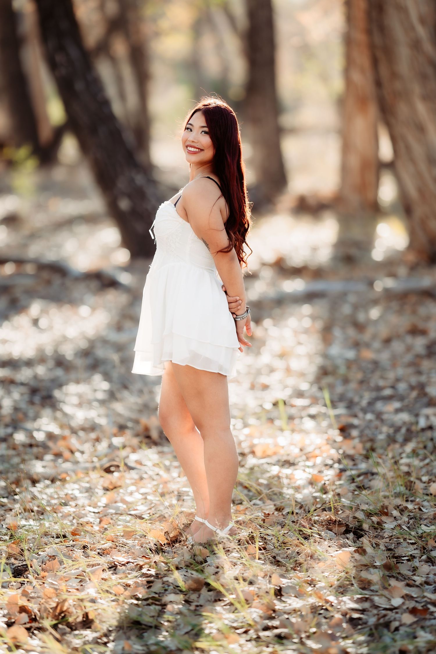 Graduate in white dress poses playfully in wooded area with fallen leaves.