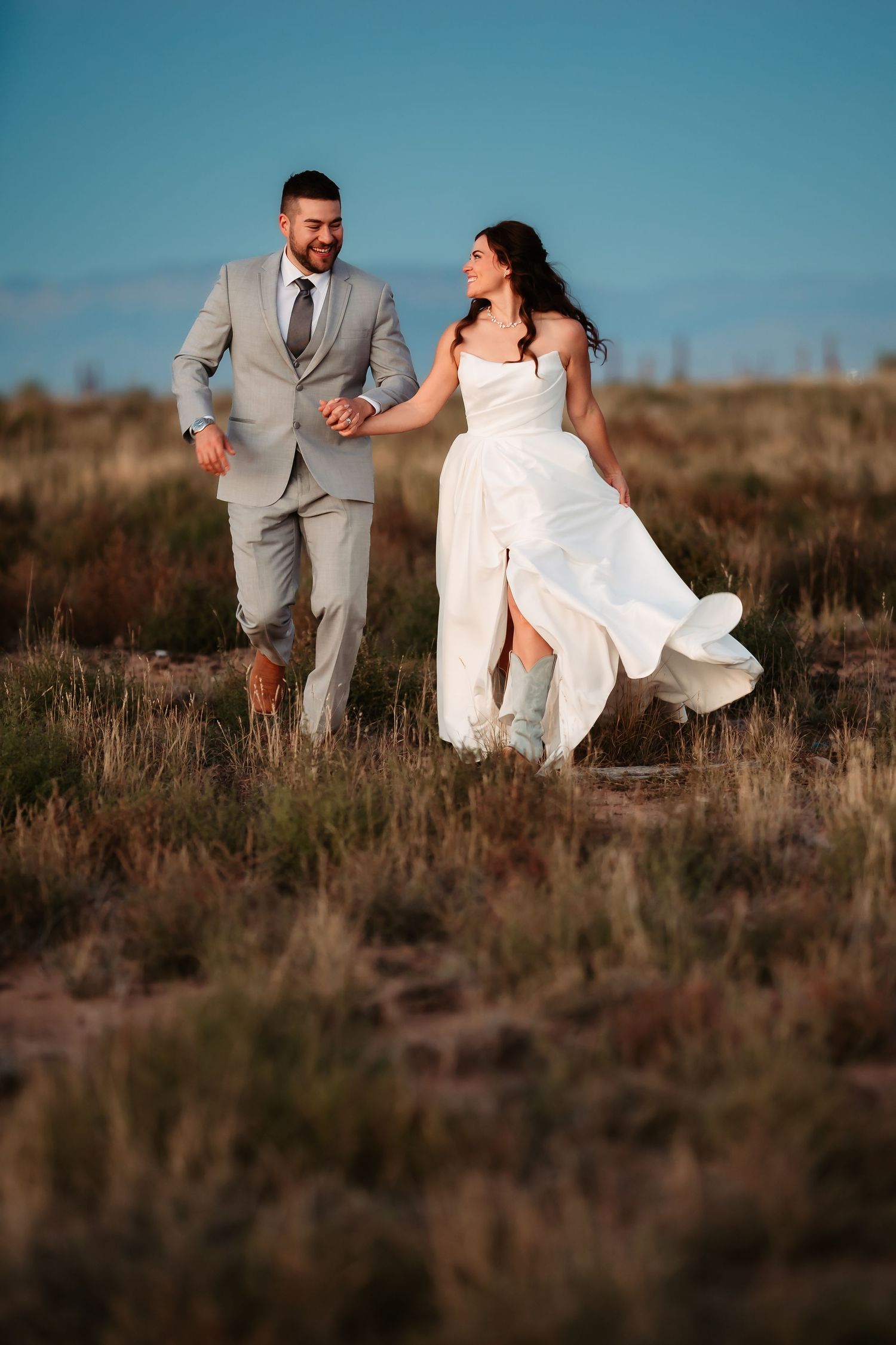 A couple in wedding attire runs playfully through a desert field at sunset.